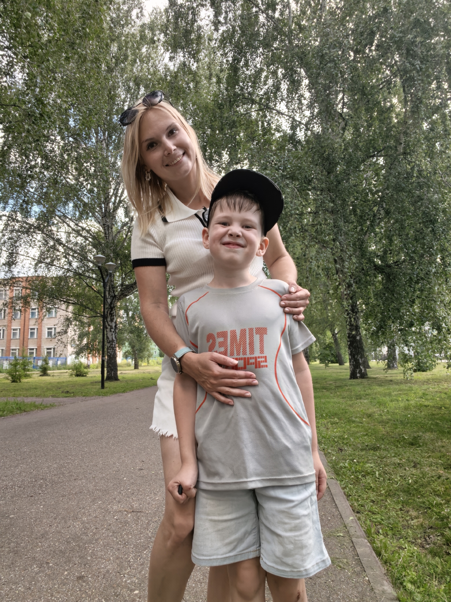 In Sterlitamak, Russia, a blond woman with sunglasses perched on her head and a young boy wearing a black baseball cap stand together on a paved walkway in a park. The woman, with a white collared shirt and a white skirt, has her arm around the boy. The boy, wearing a grey t-shirt with orange lettering and light blue shorts, smiles at the camera. Behind them, a building with multiple windows is visible, along with tall trees and green grass. A lamppost stands near the walkway. The scene appears to be a candid, outdoor portrait.