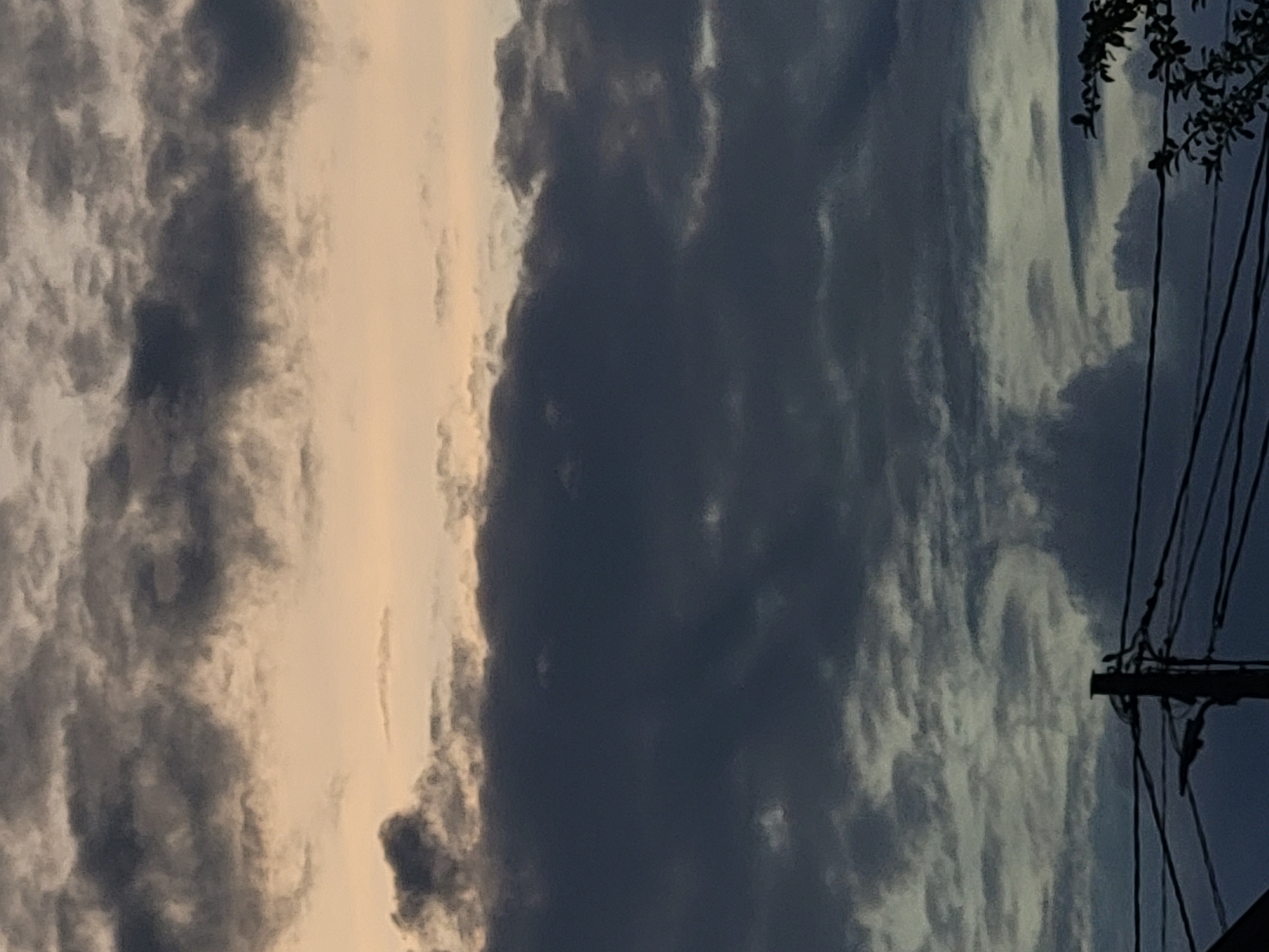 The image depicts a sky filled with heavy, dark clouds, possibly indicating an overcast day or impending rain. The lower portion of the image shows utility wires and a pole against the cloudy sky. Some tree branches are visible on the right side. The setting appears to be an urban or suburban environment, though no specific landmarks are identifiable. The overall mood is somber due to the dark clouds.