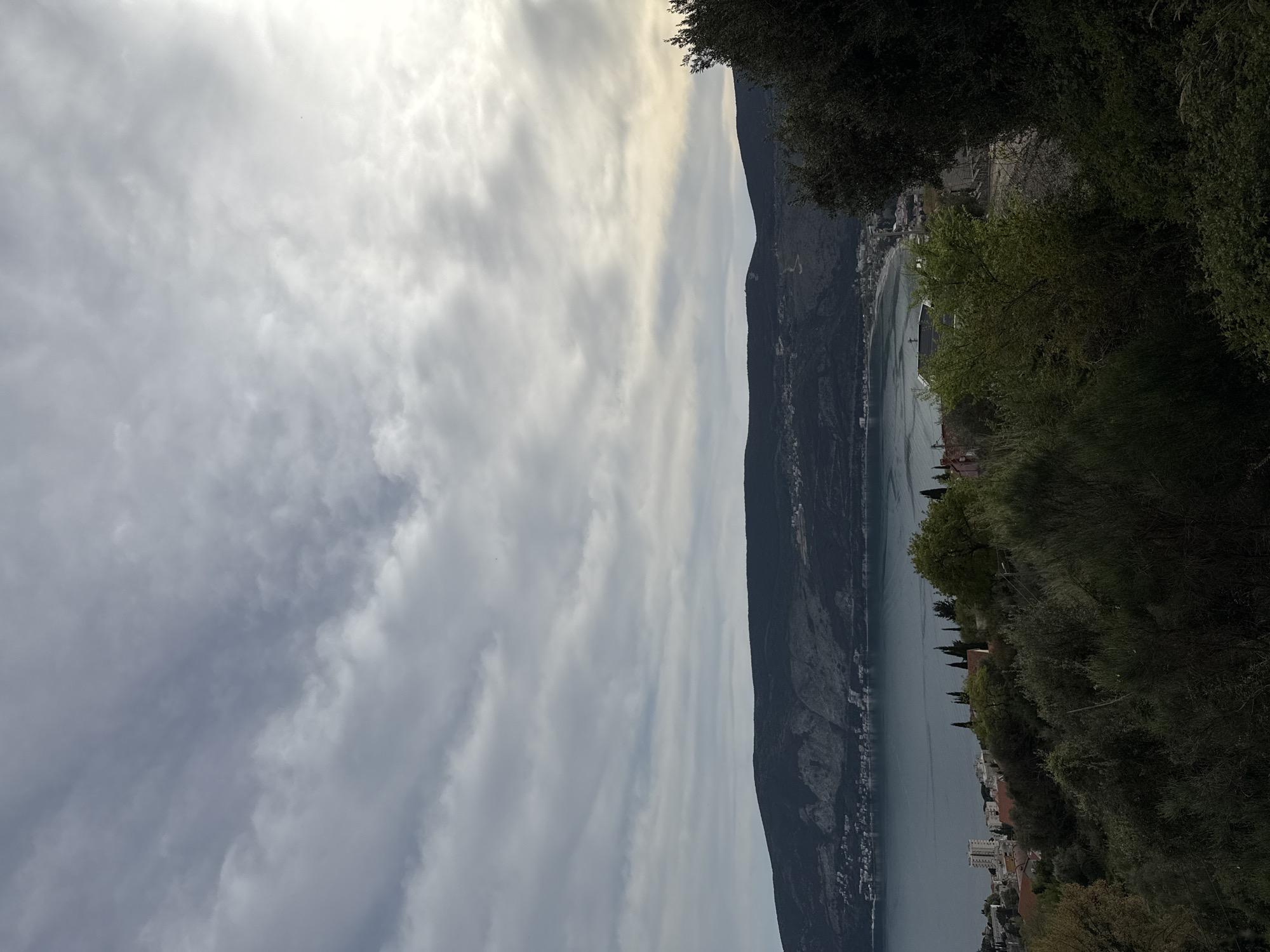 The image depicts a landscape in Trebesin, Montenegro. A long, narrow bay or inlet of water is visible, bordered by mountains on one side and a coastal town or settlement on the other. The sky is overcast with cloudy weather. Vegetation, including trees and shrubs, is present along the edges of the frame. Buildings, likely residential or commercial, are situated along the coastline. There are no people immediately visible in the image.