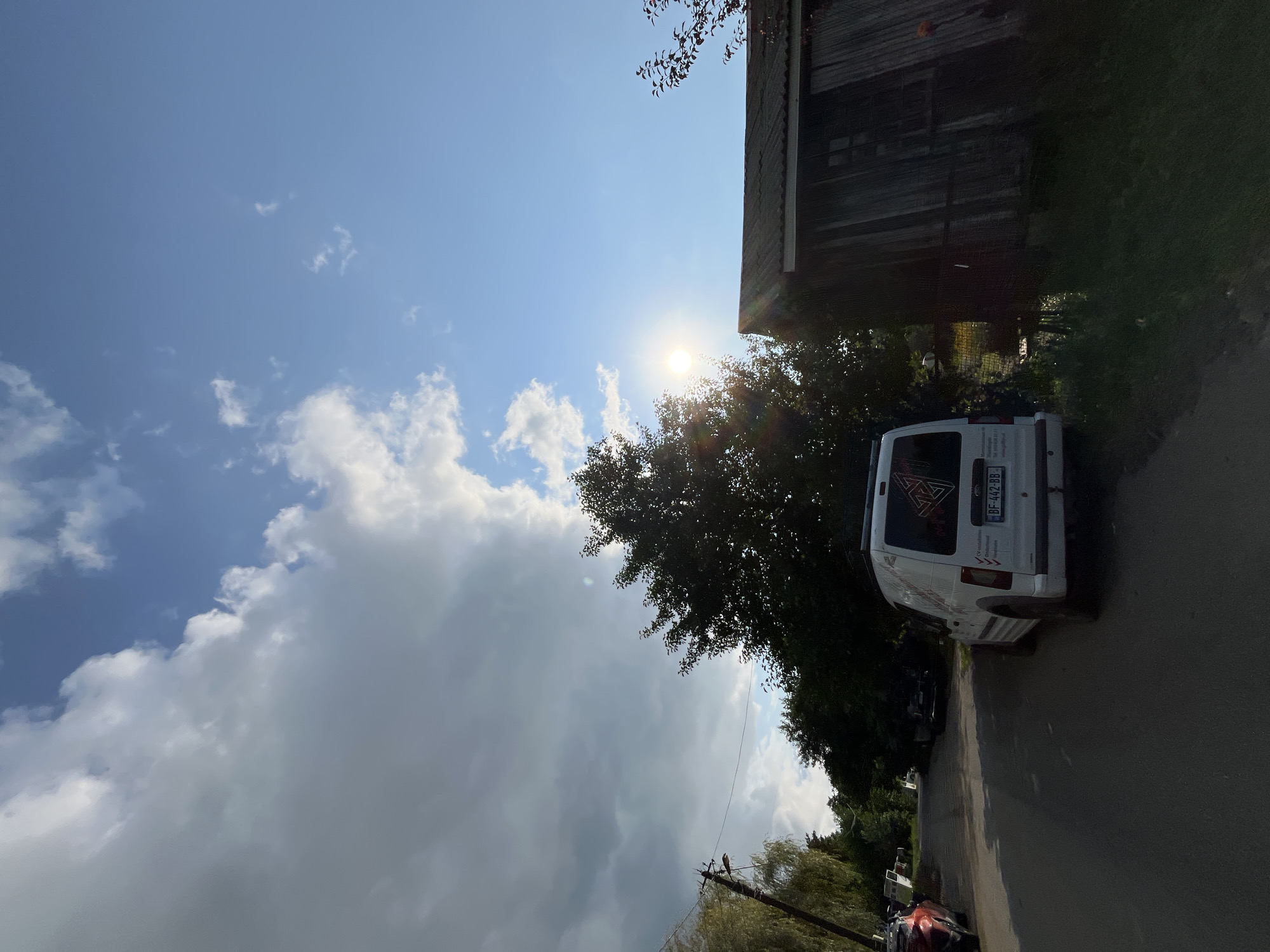 A low-angle, medium-shot captures a street scene in Ureki, Georgia, on a partially cloudy day. The sun is shining brightly, peeking through the branches of a large, leafy tree on the right side of the frame. In the foreground, a white van is parked on the side of a paved road. The van has a license plate that reads "BF 442 BB." To the right of the van, a weathered wooden structure, possibly a shed or small building, is visible. The road continues into the background, lined with trees and some other parked vehicles, suggesting a residential or semi-rural area. The sky is a vibrant blue with scattered white clouds.