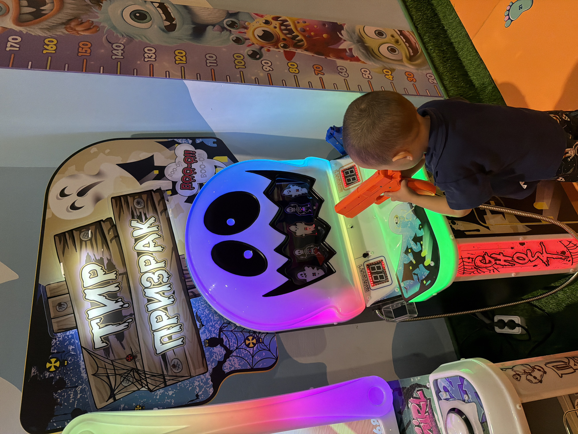 A young boy, with short dark hair and wearing a dark blue t-shirt and patterned shorts, plays an arcade game. The game features a large, illuminated ghost head with black eyes, a jagged mouth, and colorful lights that shift between blue and green. The boy is holding an orange toy gun and appears focused on the game.
The arcade machine has various spooky graphics, including cartoon monsters and cobwebs. Text on the machine reads "ТИР ПРИЗРАК" which translates to "GHOST SHOOTING GALLERY" and "ПИСТОЛЕТ 1" and "ПИСТОЛЕТ 2" (PISTOL 1 and PISTOL 2). The game also displays digital numbers, "88" and "89," on red screens.
Behind the arcade machine, a wall-mounted height chart shows numbers from 10 to 170. The chart is decorated with cartoon monster characters. The overall setting appears to be an indoor entertainment venue, possibly an amusement arcade or a children's play area. The lighting is bright and colorful, suggesting it is daytime or the venue is well-lit. The image is captured from a slightly elevated angle, looking down at the boy and the game.