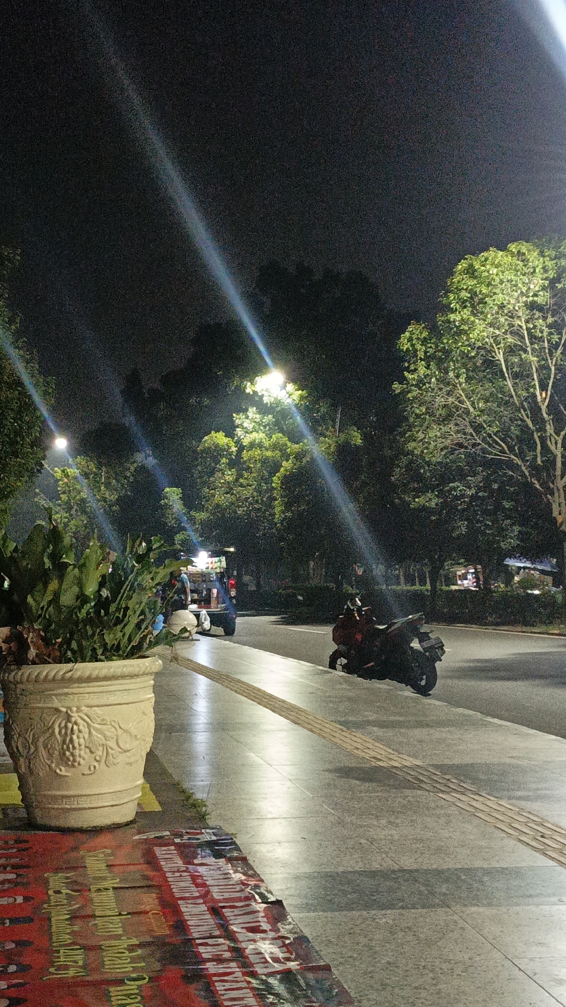 This nighttime photograph captures a scene on a street in Tigaraksa, Indonesia. In the foreground, a large, ornate beige planter holds lush green foliage. To the left, a red banner with faded white and yellow text is laid out on the ground. The sidewalk is illuminated by artificial lights, casting long reflections.
Further down the street, a food cart with people around it is visible. A dark-colored motorcycle is parked on the right side of the road, facing away from the viewer. Tall trees with illuminated canopies line the background, creating a dark, atmospheric setting punctuated by light beams from streetlights. The sky is dark and overcast.