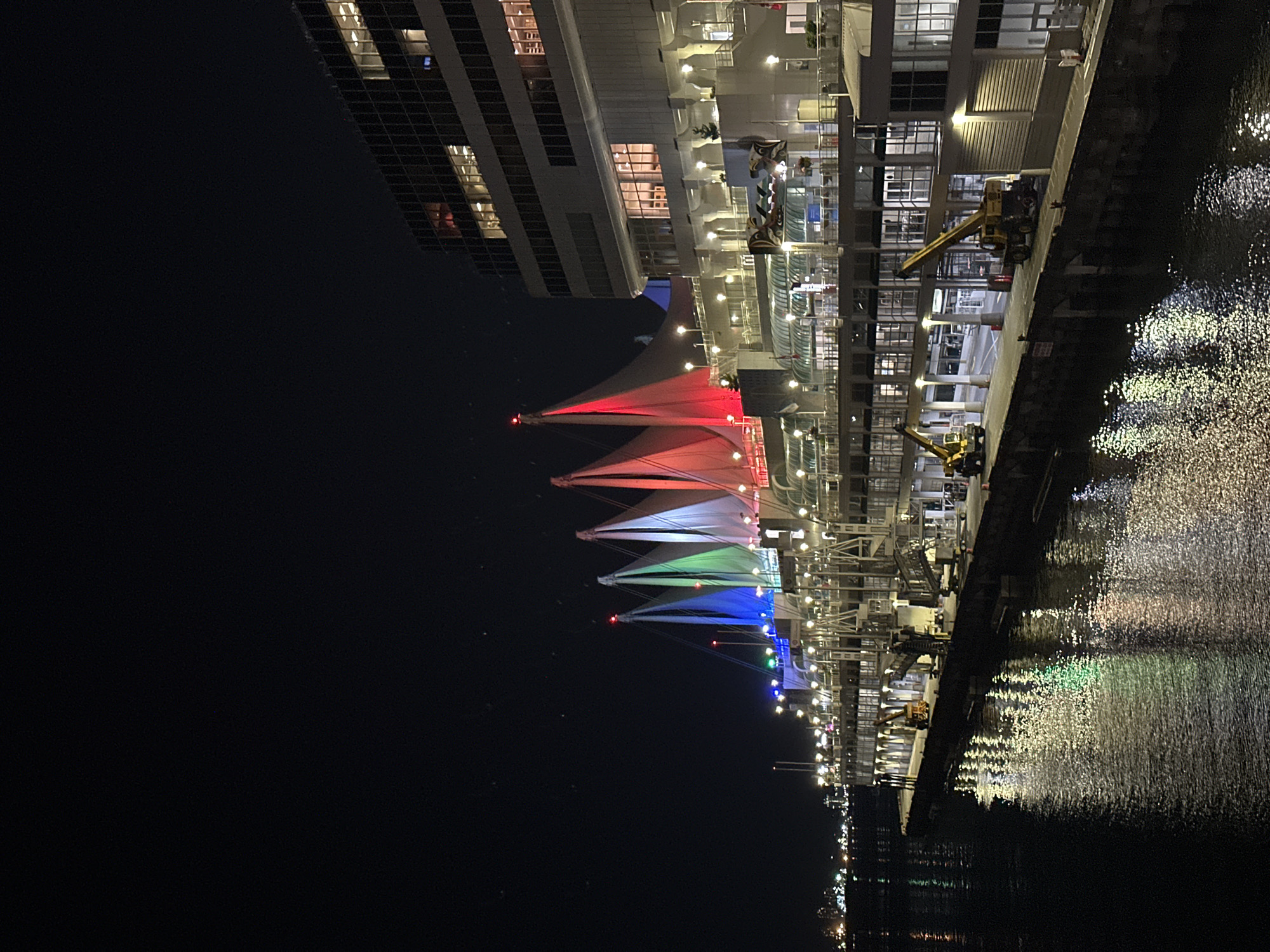 The image shows a nighttime view of a waterfront building with sail-like structures illuminated in red, green, and blue lights. The building has multiple levels of windows and balconies, with lights on inside some of the rooms. A modern building with glass facades rises behind the waterfront structure. Two yellow construction cranes are parked on the dock. The water in the foreground reflects the colorful lights from the building and the sky appears dark with a few faint stars. The overall scene depicts a modern urban waterfront at night.