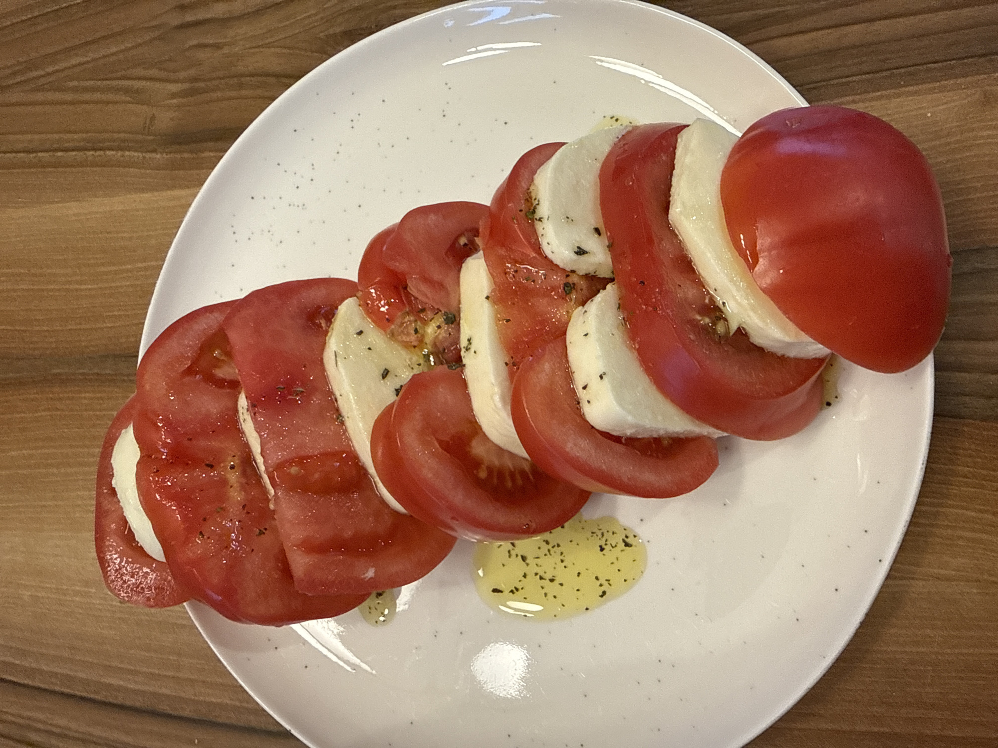 A close-up, high-angle shot shows a Caprese salad arranged on a white, speckled plate. The salad consists of alternating slices of ripe red tomatoes and fresh white mozzarella cheese, stacked in a gently curved line. Drizzled over the salad is a golden olive oil, sprinkled with dried herbs, likely basil or oregano, and black pepper. The plate rests on a warm-toned wooden surface, with a shallow depth of field blurring the background. The image was taken in the city of Plovdiv, Bulgaria.