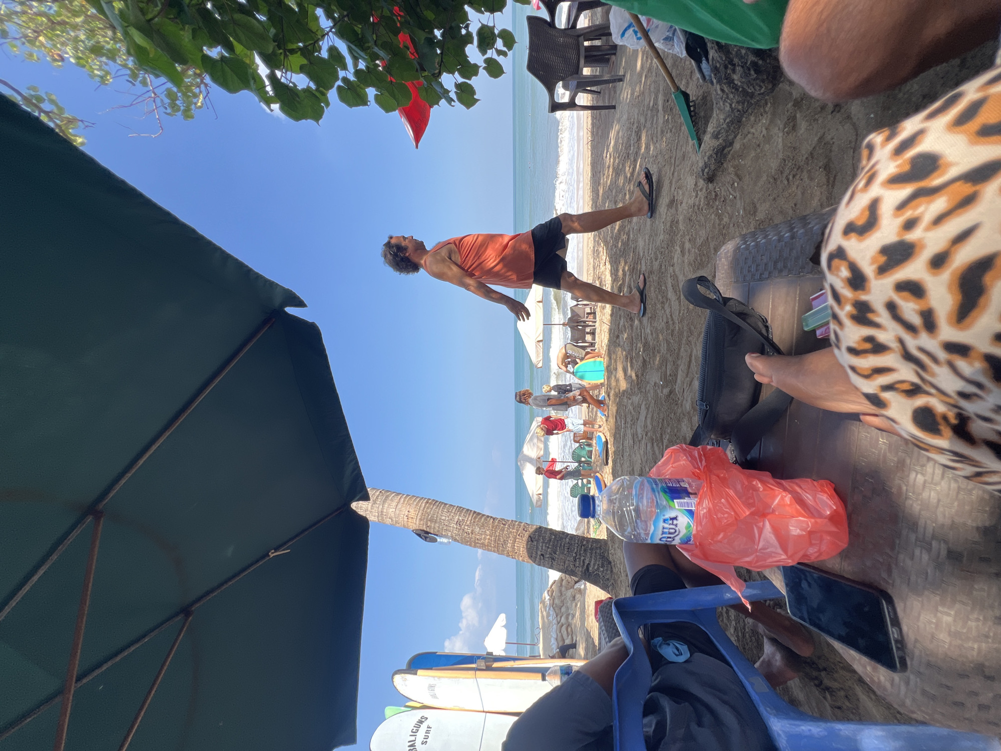 A group of people are on a sunny beach in Kuta, Indonesia, with the ocean visible in the background. In the foreground, a person sits in a blue plastic chair under a large dark umbrella, with a cell phone and a bottle of Aqua water in a red plastic bag on a brown woven table in front of them. Further back, a man in an orange tank top and black shorts walks along the sand. Several surfboards are propped upright near the left side of the image. In the middle ground, a few beach umbrellas are set up, and other beachgoers can be seen walking and relaxing. The sand is light brown, and the water is blue with white waves. A palm tree stands between the foreground and the middle ground.