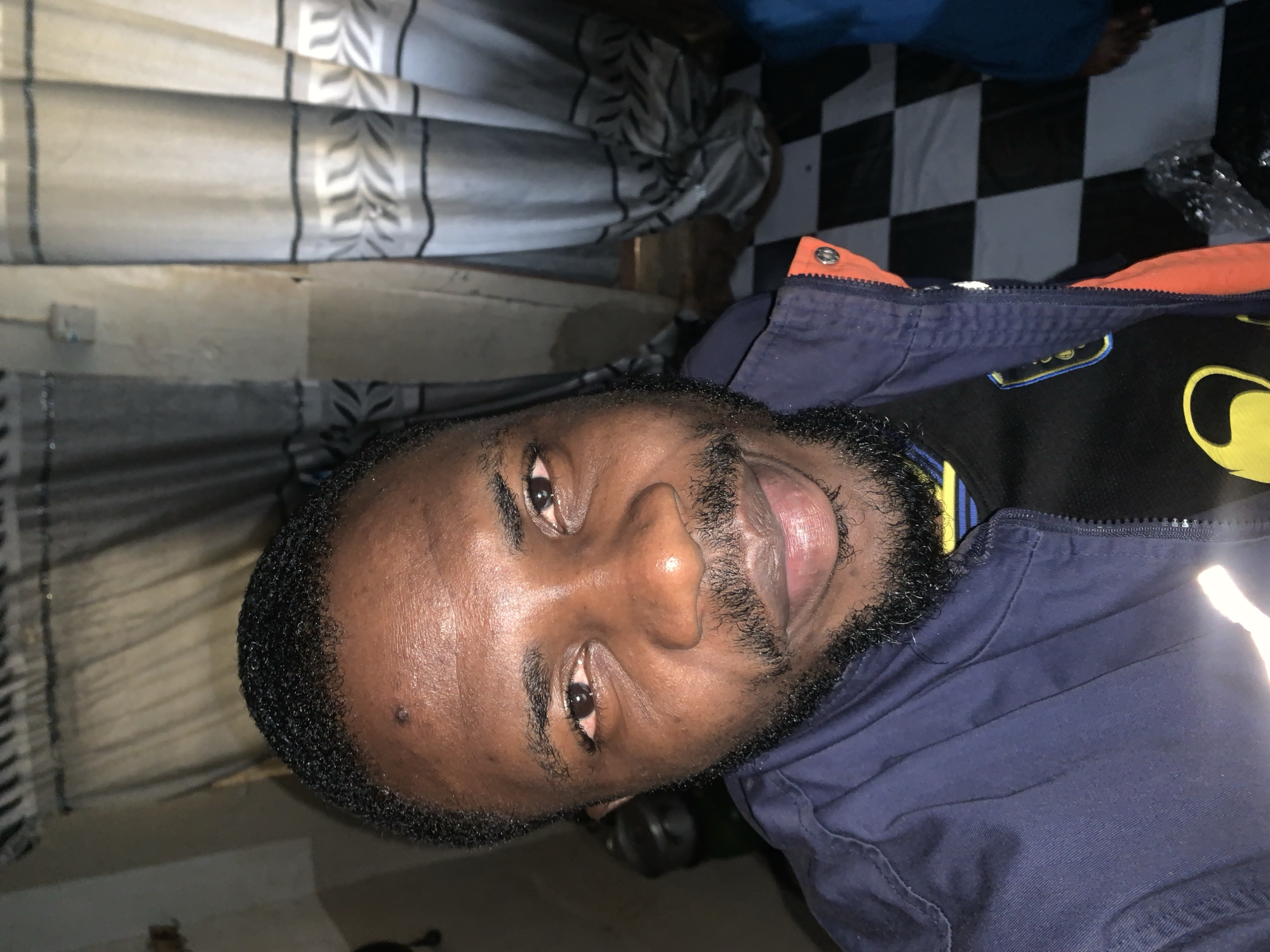 In New Stadium, Nigeria, a medium close-up selfie shows a Black man facing the camera, with his head and shoulders filling most of the frame. He has short, curly black hair, a full beard, and is smiling with his lips slightly parted. His skin is dark brown, and he has some redness around his eyes. He is wearing a blue jacket over a black shirt with yellow and blue accents. The background is dimly lit, with a patterned curtain visible on the right and what appears to be a tiled floor with a black and white checkered pattern.