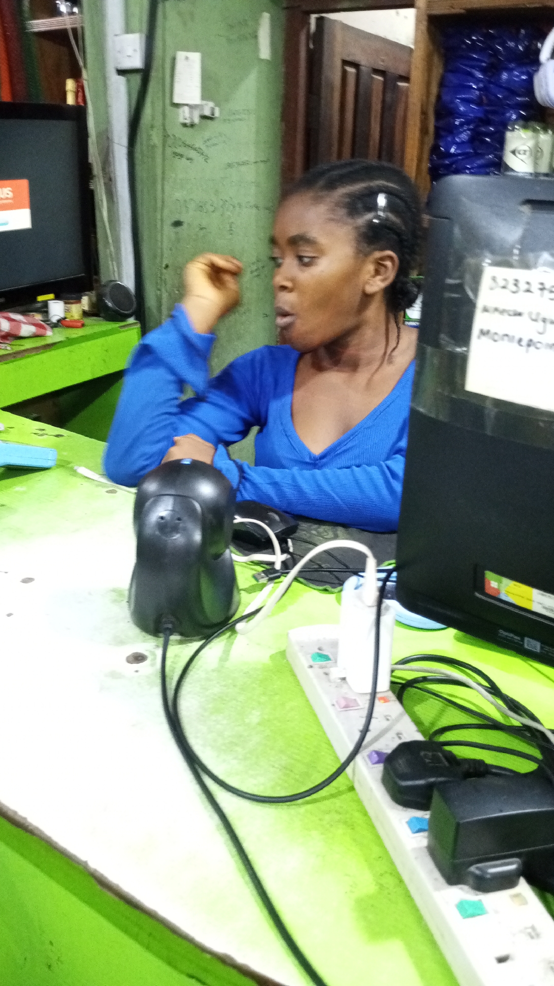An indoor scene captures a young woman working at what appears to be a service counter or small shop in Adankolo, Nigeria. The woman, with dark skin and braided hair styled in cornrows, wears a blue long-sleeved top with a V-neck. She is seated, leaning slightly forward, with her right hand near her chin and her mouth slightly open, suggesting she is speaking or listening intently, conveying a sense of engagement.
The workstation features a bright green desk or counter. Prominently in the foreground is a black barcode scanner. To the right, a black computer monitor is partially visible, with a piece of white paper taped to its side. This paper displays handwritten text: "323275", "Annan Ugu", and "Moneypoint," indicating a potential payment or financial service. A tangled array of black and white cables connects various electronic devices, including a white power strip with multiple colored outlets, many of which are occupied by plugs.
To the left, another screen or television monitor is visible, displaying a partial logo with the letters "US." A small black speaker sits next to it. The wall behind the woman is light green and shows some faint handwritten marks, as well as a white wall socket. A dark wooden door is visible in the background, and above it, a shelf holds several blue plastic bags. The overall setting suggests a busy, functional workspace within a local business.