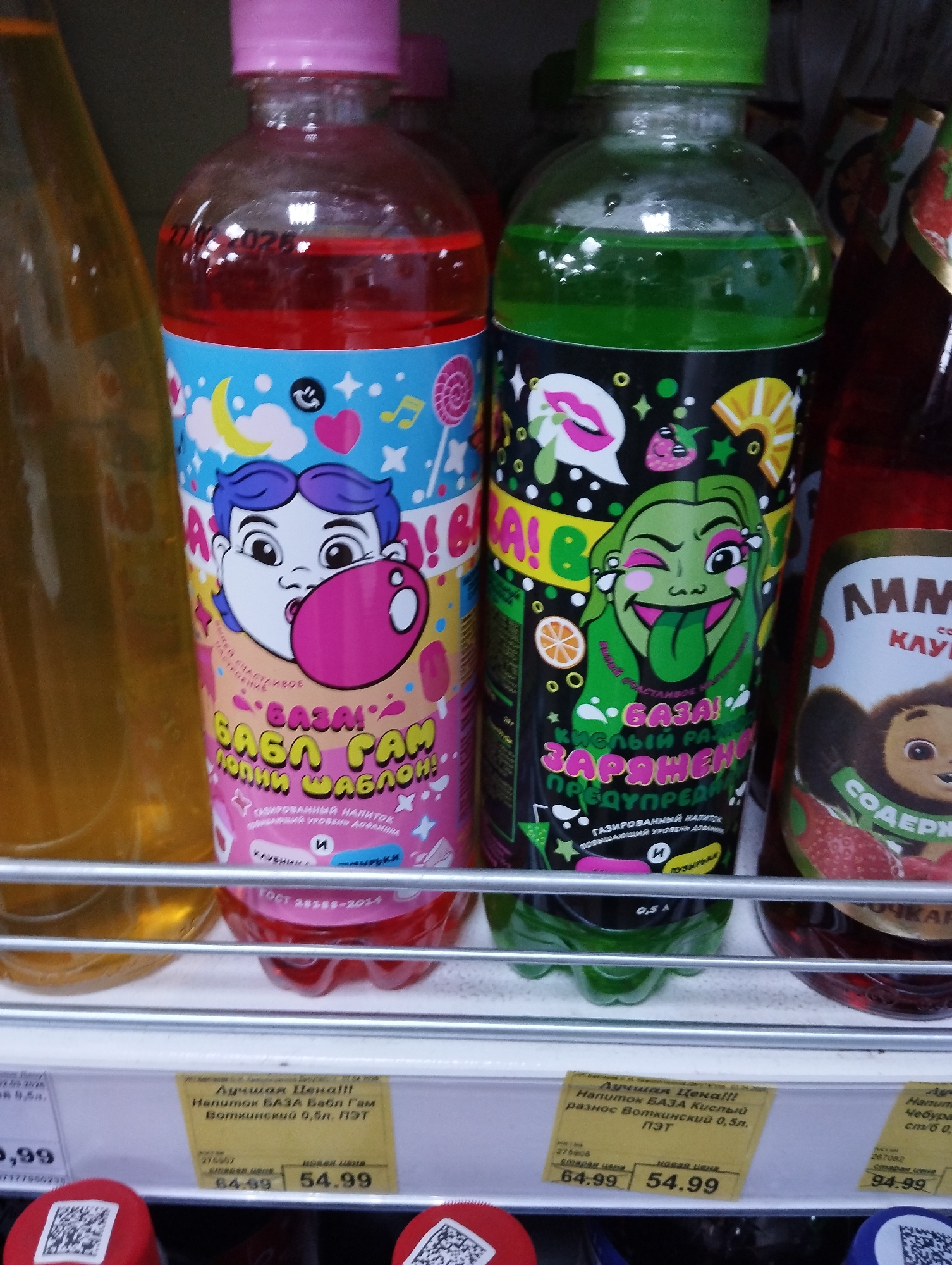 The image shows two bottles of a carbonated beverage called "Baza" on a store shelf. The bottles are made of clear plastic and have colorful labels. The bottle on the left has a pink cap and a label featuring a cartoon character blowing a pink bubble. The bottle on the right has a green cap and a label featuring a green monster-like character winking. Below the bottles, a price tag indicates that the beverage is sold at a reduced price of 54.99. The text on the label of the left bottle says "Baza! Babl Gam Topolniy Shablon! Gazirovanniy Napitok", which translates to "Baza! Bubble Gum Topolniy Shablon! Carbonated Drink". The text on the right label says "Baza! Kisliy Raznos Zaryazhen Predoprezhden! Gazirovanniy Napitok", which translates to "Baza! Sour variety Charged Warn! Carbonated Drink". The description on the yellow label says that the drink is from the company Volzhskiy. The location of the image is in the city of Krasnogorskoye, Russia.