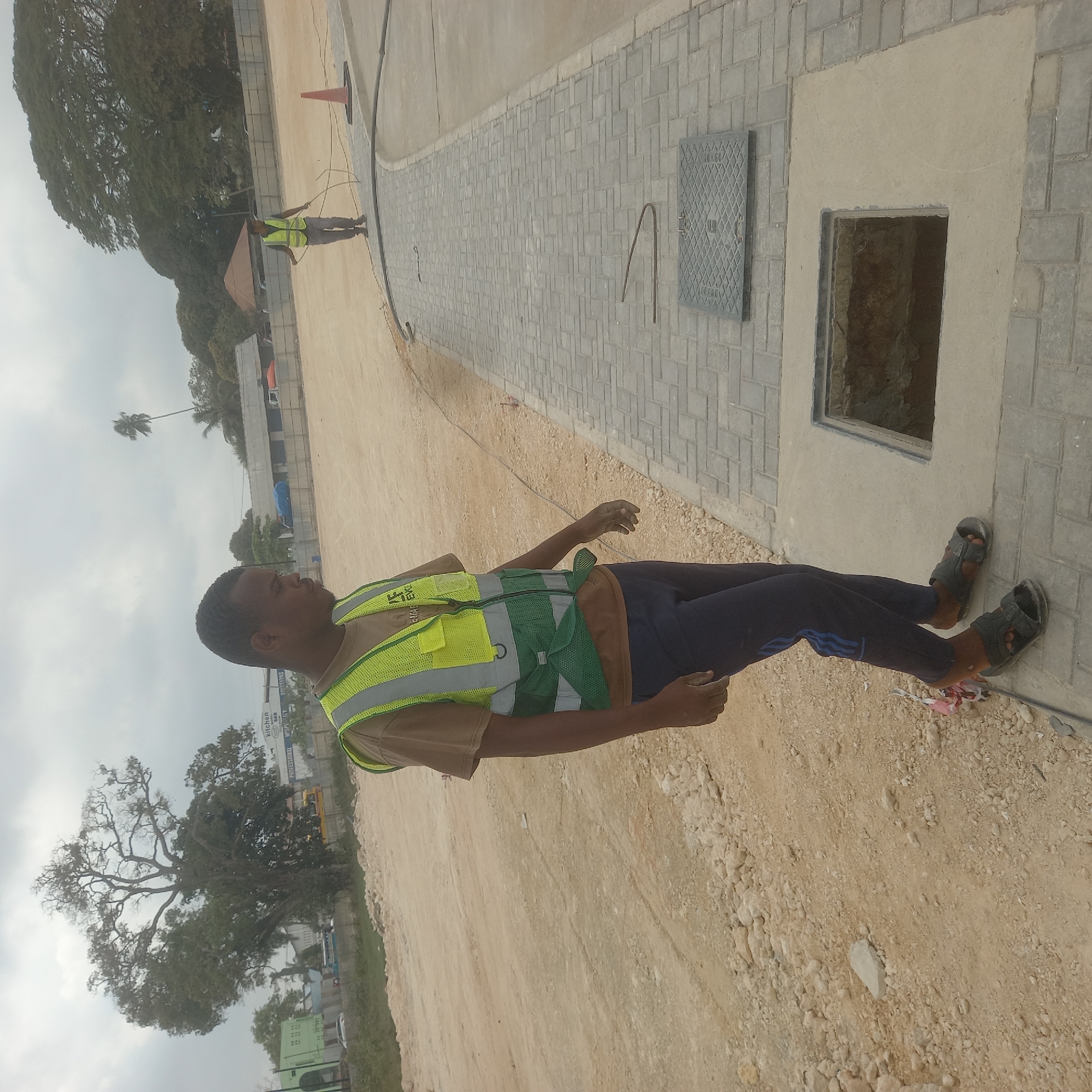 In the city of Bububu, Tanzania, a young man wearing a tan t-shirt and a bright green and yellow safety vest stands on a dusty, unpaved construction site. He is wearing dark blue track pants with a blue stripe and black sandals. His gaze is directed downwards, and his hands are visible in front of him. To his right, a paved walkway made of interlocking grey bricks leads away from the viewer. A rectangular opening, revealing dark earth below, is set into the concrete next to the walkway. Further down the walkway, a man in a similar safety vest is visible in the distance, holding a coiled wire and standing near an orange traffic cone. The background consists of trees, some with sparse foliage, and a cloudy sky. Buildings, including one with a green facade, are visible in the distance. The overall scene depicts an active construction area.