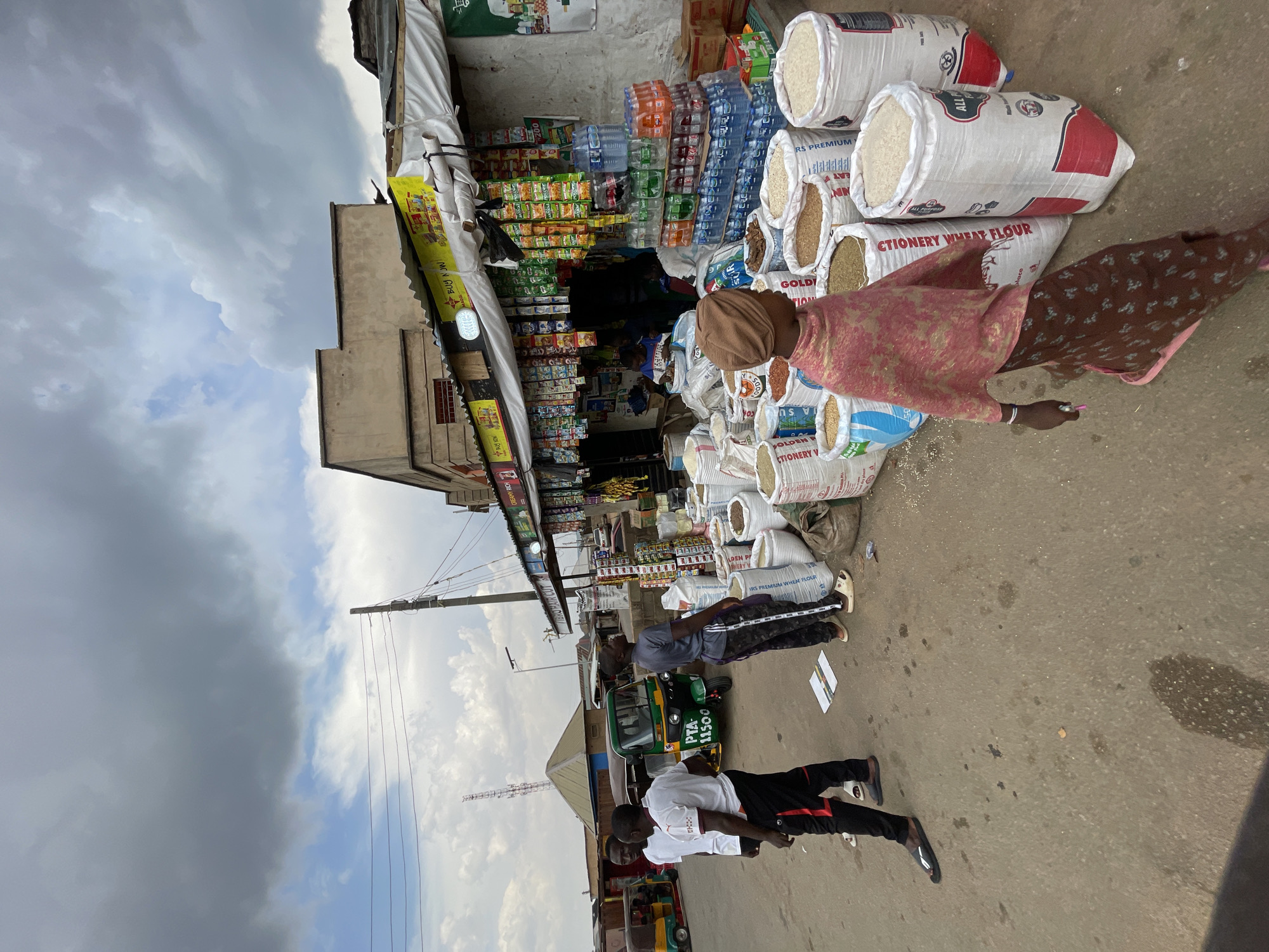 The image depicts a street scene in Jos, Nigeria. A store with an awning displays a variety of goods, including stacks of water bottles, snacks, and large bags of flour and grains. Several people are present: a woman in traditional attire stands near the bags, and a few men are visible further down the street next to a green three-wheeled vehicle (a "Keke"). The sky is cloudy. The street itself is paved, and the overall impression is of a bustling commercial area.
