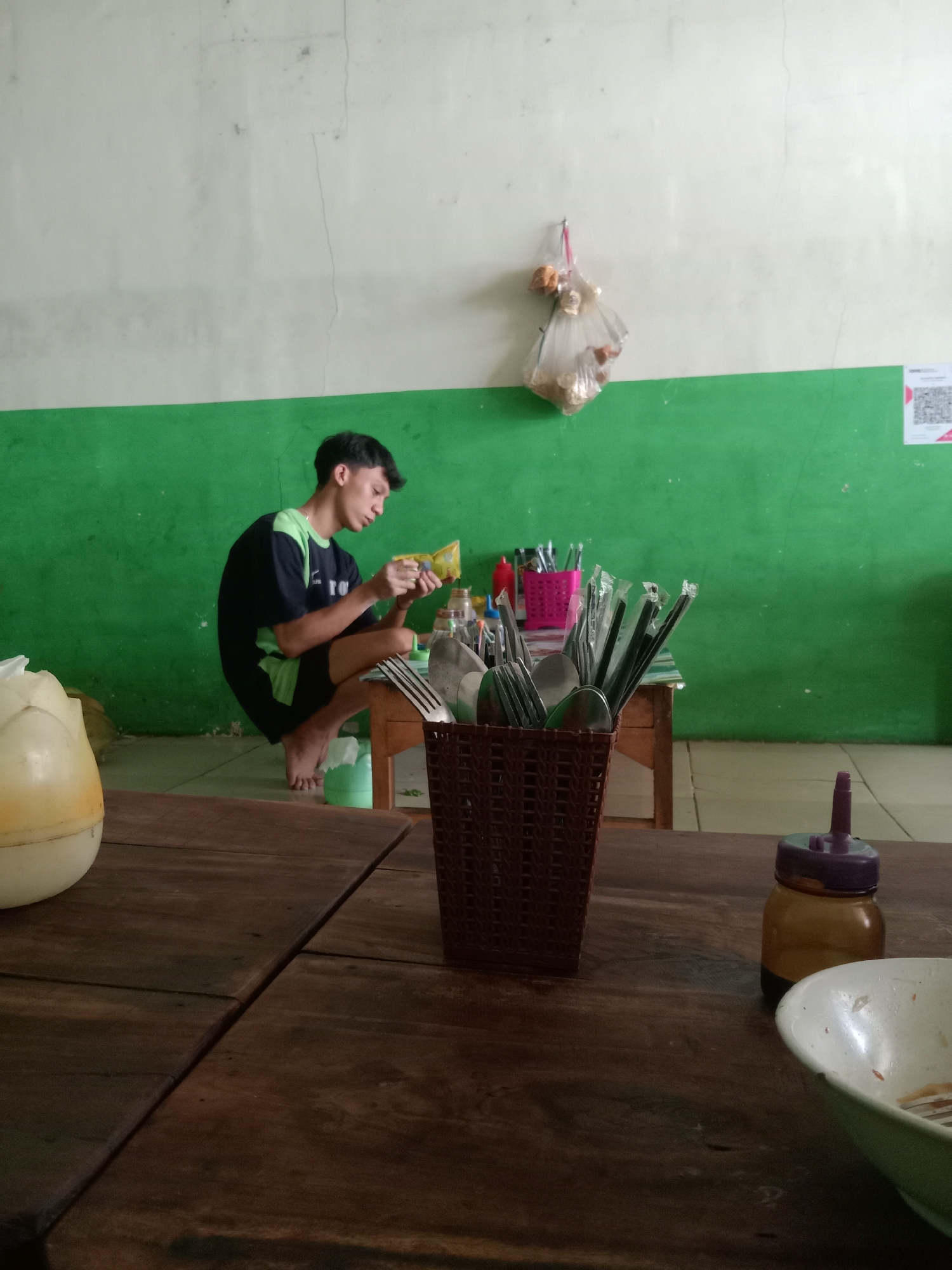 The image depicts an indoor scene, likely a simple eatery, in Sugihwaras, Indonesia. In the mid-ground, a young man, wearing a dark t-shirt with neon green accents and dark shorts, is crouched on the floor. He is holding a yellow packet and appears to be preparing or examining its contents. He is barefoot.
In the background, a green-painted wall stretches across the scene, meeting a white-painted upper wall. A bag of food items is hanging on a hook on the white wall. A small wooden table is positioned in front of the green wall, on which a pink plastic container holds utensils, and a brown woven basket contains more cutlery, including forks and spoons, some of which are individually wrapped in plastic. Bottles of condiments are also visible on the table.
The foreground is dominated by a dark wooden table, showing signs of wear and tear. A light-colored, possibly plastic, container with a floral-like design is partially visible on the left side of the table. On the right side of the foreground table, a small amber glass bottle with a purple plastic dispenser top is present, and a white bowl with some residue is also visible. The lighting suggests an ambient indoor setting, possibly during daytime.