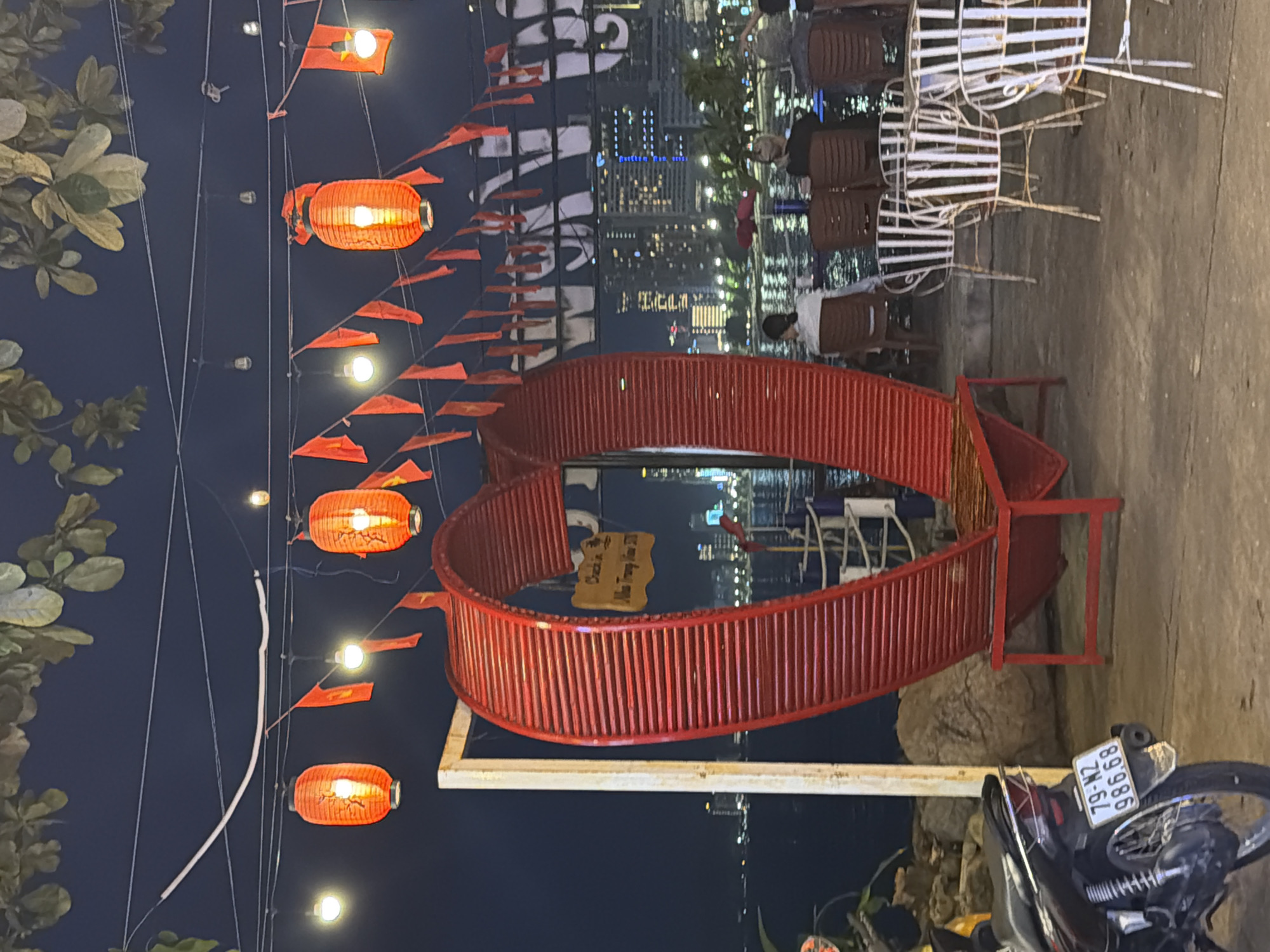 A nighttime scene unfolds in Nha Trang, Vietnam, characterized by a waterfront setting and festive decorations. In the foreground, a large, red, heart-shaped bench is prominently displayed, constructed from slatted metal. To the left, a black motorcycle is parked, displaying a license plate that reads "79-M2 986.68". Several white metal chairs with slatted seats are arranged in the mid-ground.
The backdrop features a body of water with distant city lights reflecting on its surface. A skyline of multi-story buildings is visible across the water. Overhead, red paper lanterns and small orange flags are strung across wires, illuminated and contributing to the ambiance. Several individuals are seated in chairs in the mid-ground, facing the water, their backs mostly to the camera. The overall atmosphere suggests an outdoor seating area or a viewing point in a coastal urban environment.
