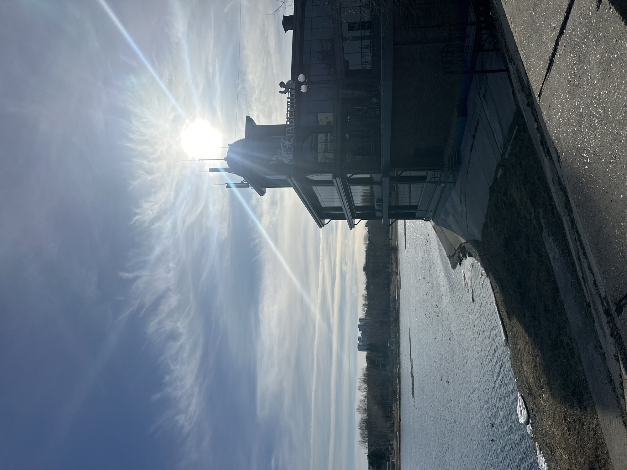 This media file presents an outdoor landscape featuring a prominent building, a body of water, and a bright sky, likely captured in Irkutsk, Russia.
The image is dominated by the intense sun, positioned high in the upper left, casting a strong sunburst effect and lens flare across the frame. The sky is a light blue with wispy, high-altitude clouds, some appearing as long, straight contrails, suggesting a clear but cool day.
On the left, a multi-story building with dark facades and large windows stands beside a paved path. A notable feature on the building's front is a sign with Cyrillic text that reads "ГРАФЪ КАЛАШНИКОВЪ" (Graf Kalashnikov), indicating it is a commercial establishment, possibly a restaurant or café. A small, decorative figure of a man is visible on the roof near the sign, and streetlights are mounted below it.
To the right of the building, a paved area transitions to a grassy or dirt embankment that slopes down to a wide body of water, likely a river or lake. The water's surface shows ripples and reflections, and along the immediate shoreline, patches of snow or ice are visible on the ground, hinting at a colder season such as late winter or early spring.
Across the water, a distant treeline of bare trees marks the horizon, with some urban structures or taller buildings faintly visible beyond, consistent with an urban or semi-urban setting. The time of day appears to be midday or early afternoon, given the high position and intensity of the sun. There is no visible human activity, creating a serene and quiet atmosphere.