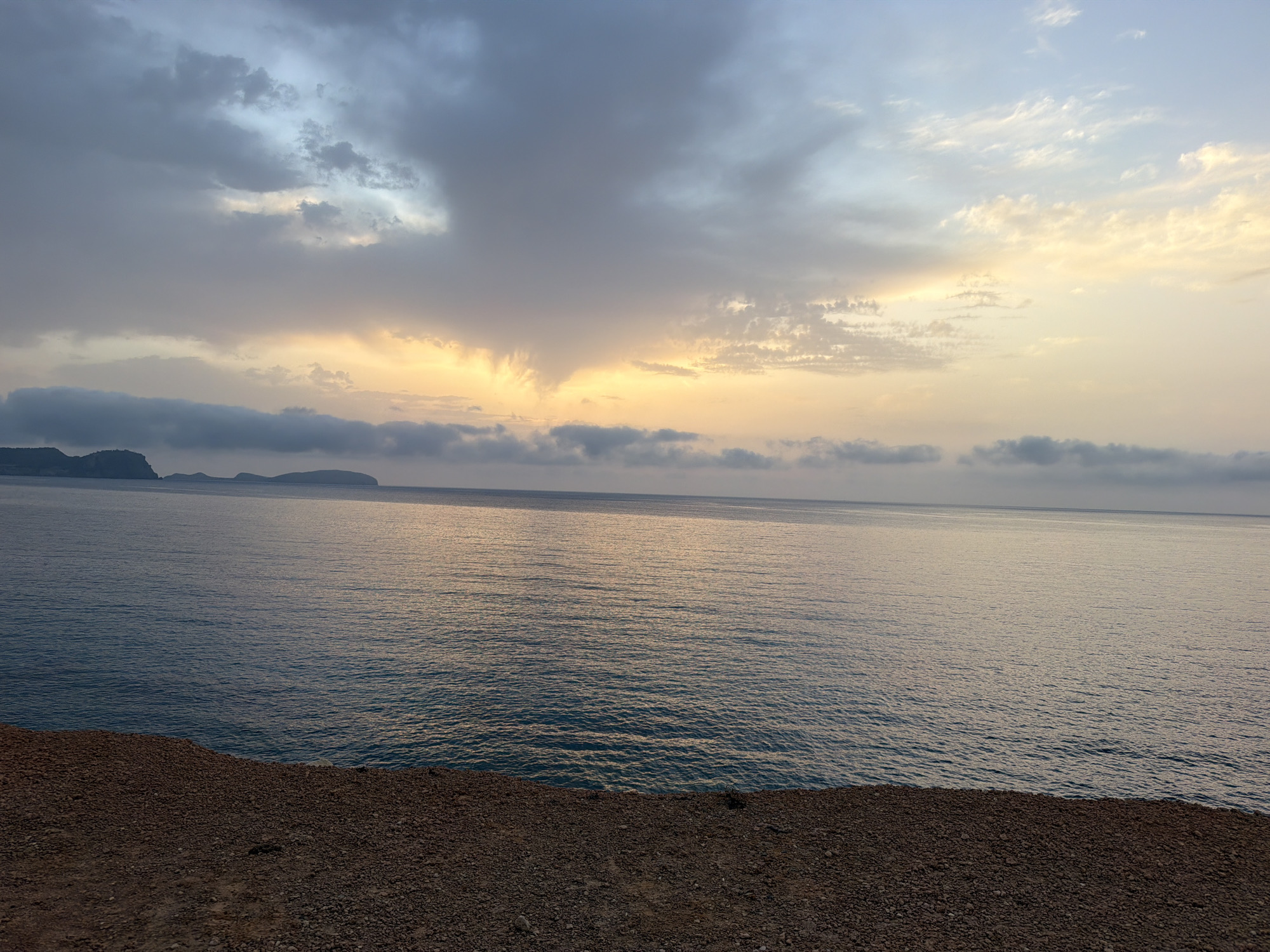 The image depicts a coastal landscape at dusk, with the sea in the foreground and islands in the distance under a cloudy sky. The sky is a mix of gray, blue, and hints of yellow and orange where the sun is setting or has recently set. The sea is a deep blue with gentle ripples reflecting the sky's colors. The foreground is a rocky, reddish-brown cliff or embankment. In the far distance, silhouetted against the horizon, are several low-lying islands. The scene suggests a peaceful, natural setting on the coast of Es Canar, Spain. There are no people or man-made objects visible in the image.
