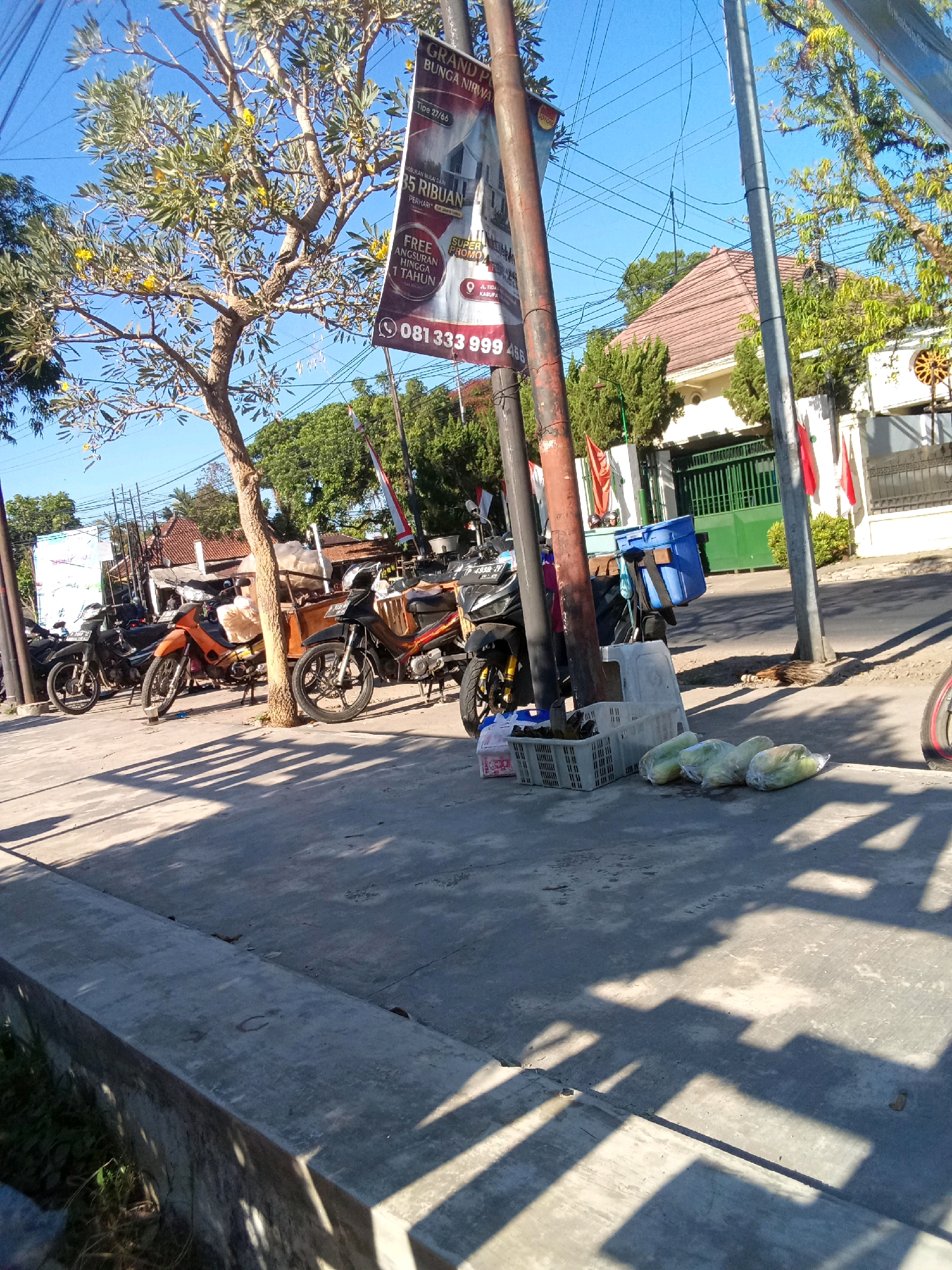 A street scene in Jember, Indonesia, shows several motorcycles parked along a sidewalk next to a road. The motorcycles appear to be of various models and colors. Some are leaning on their kickstands, while others are supported by a central stand. A tree with yellow flowers is partially visible on the left side of the image, casting shadows on the sidewalk. Several white plastic crates and a blue plastic container are placed on the sidewalk near the motorcycles. In the background, a building with a red-tiled roof and a green gate is visible, along with trees and utility poles. A large banner advertising "GRAND P BUNGA NIRWA" is hanging from a pole. Several red and white flags are also displayed. The overall lighting suggests it is daytime, with a clear blue sky. There are no people clearly visible in the image, though there may be individuals obscured by the motorcycles or out of frame.