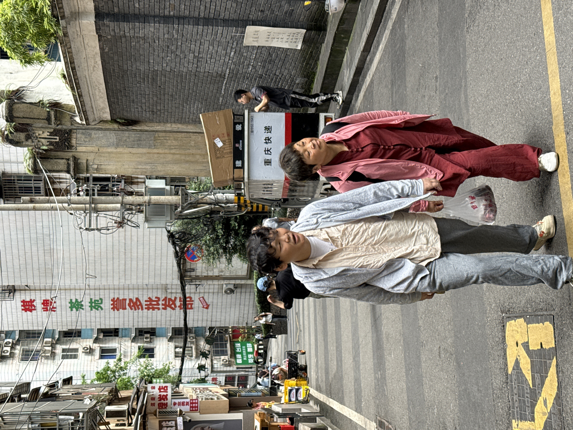 The image shows a street scene in the Yuzhong District of China. In the foreground, two people are walking. One is wearing a light gray jacket over a beige shirt and gray pants. The other is wearing a red outfit with a pink jacket. Behind them is a parked truck with "重庆快递" (Chongqing Express) printed on the side. A man is seen near the truck. In the background are buildings with Chinese signs, including "棋牌 茶苑誉多批发部" (Chess and Card Room Yudu Wholesale Department) and "理发店" (barbershop). There are air conditioning units on the building exteriors. The street also features tables, chairs, and various objects. A yellow line is painted on the pavement.