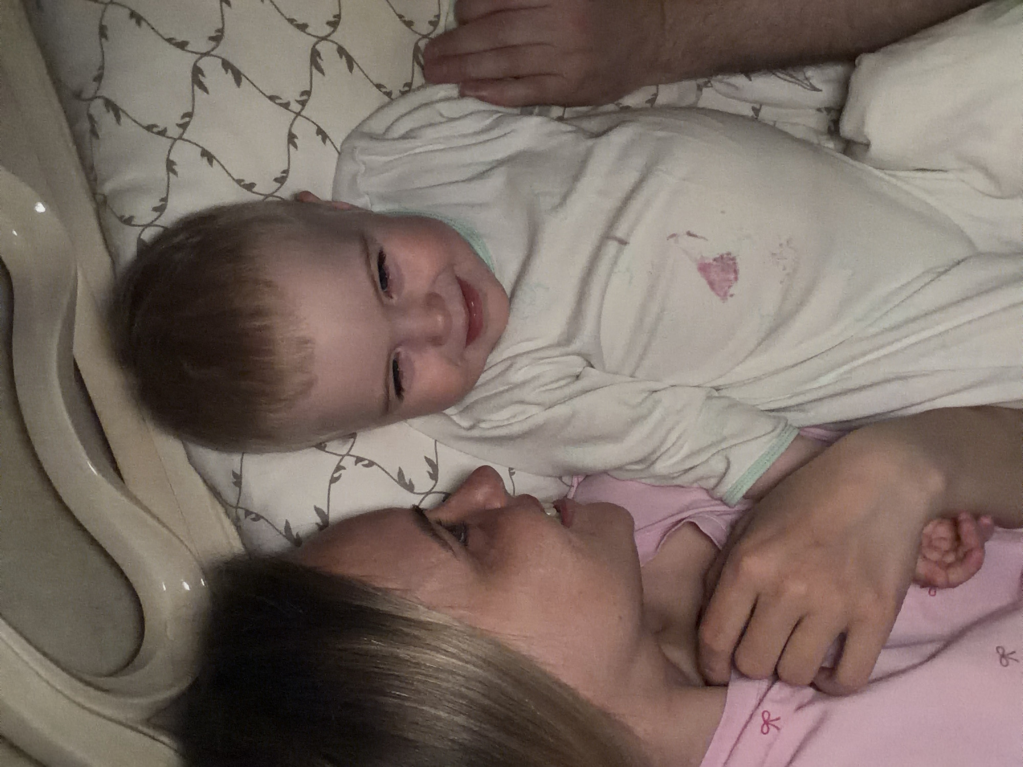 A woman and a baby are lying on a bed. The baby is on top, wearing a white onesie and smiling. The woman is lying below, looking up at the baby. A man's hand is partially visible above the baby. The bed has a patterned pillow and white sheets. The scene may be taking place in a home in Votkinsk, Russia, but the location cannot be definitively confirmed from the image alone.