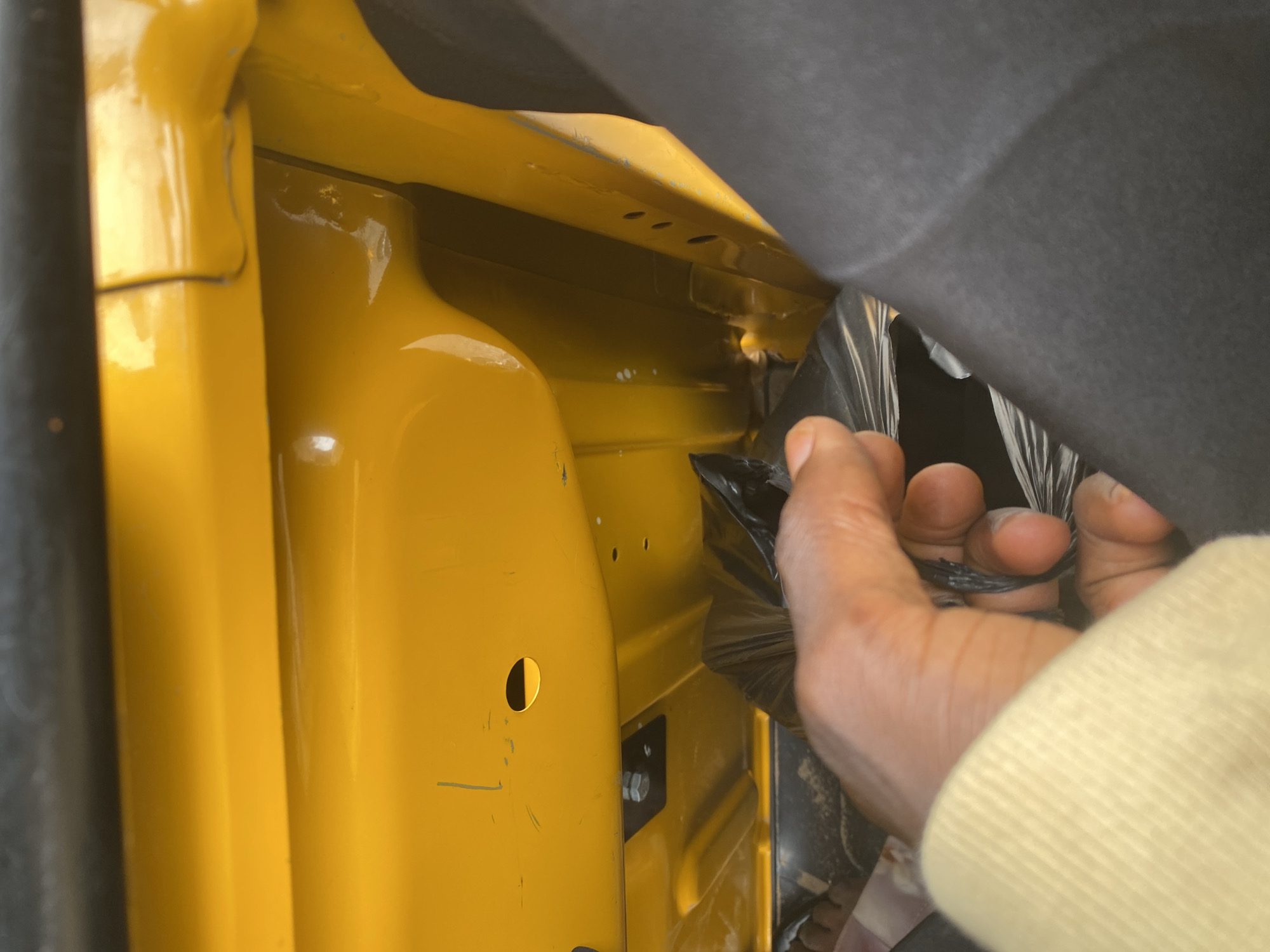 A hand wearing a light yellow sleeve is holding a black plastic bag inside a yellow vehicle. The vehicle's interior is predominantly yellow, with visible metal components and some scuff marks. There is a hexagonal bolt secured to a black plate on the vehicle's frame. A circular hole is also visible on the yellow paneling. The context suggests a location within Jalingo, Nigeria, potentially related to transportation or vehicle maintenance.