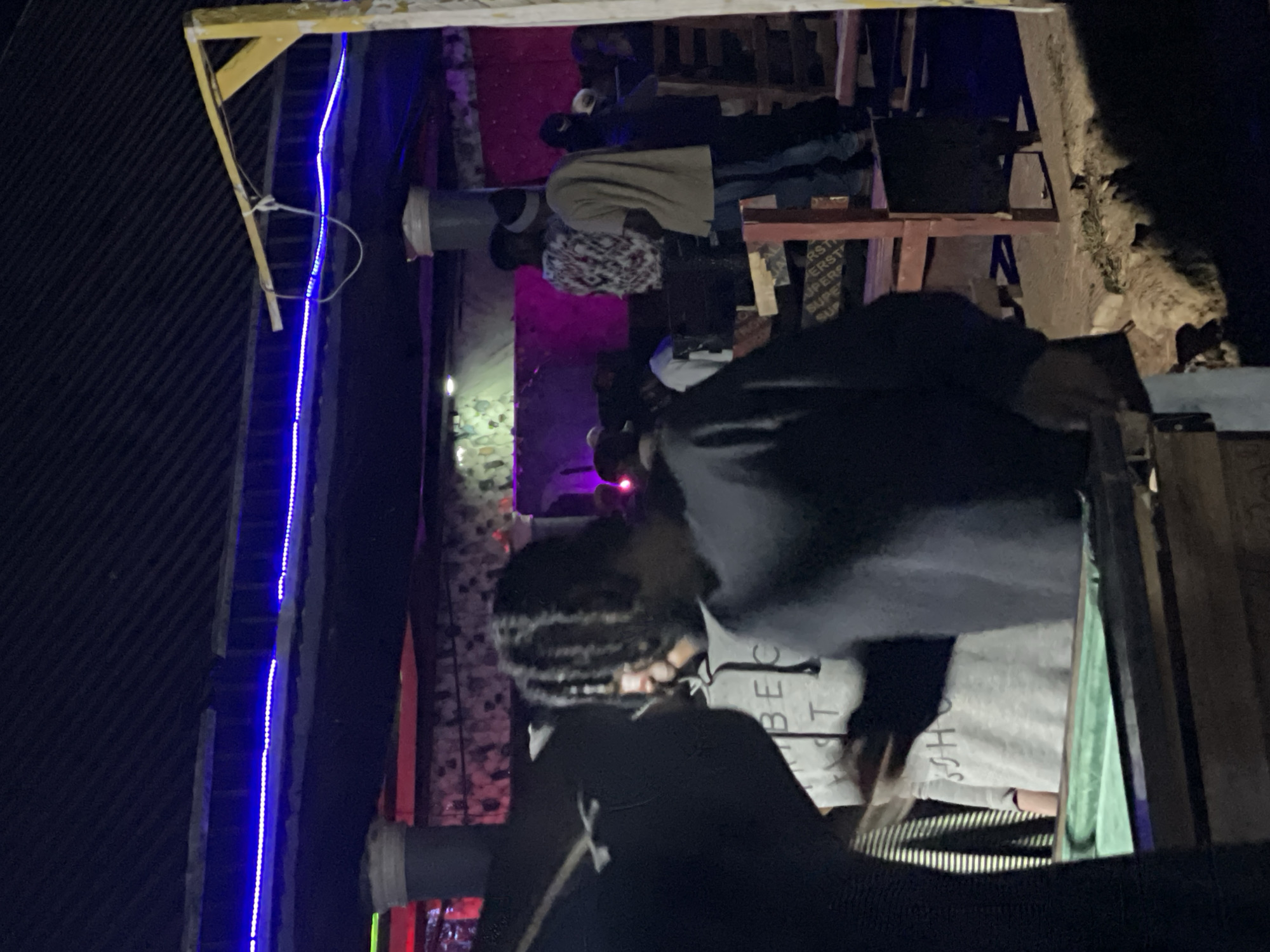 The image depicts an outdoor nighttime gathering, possibly in Durumi, Nigeria, with a focus on a group of people socializing. In the foreground, a person with braided hair is facing away from the camera, wearing a dark long-sleeved shirt. To their left, another individual is partially visible, wearing a gray hoodie with "BEC T" and "SOHO" printed on it, positioned near a green-topped table, possibly a pool table. The background shows several other individuals, some standing and others seated, amidst a dimly lit environment illuminated by blue and red LED lights. The setting appears to be a casual outdoor space with wooden seating and a corrugated metal roof structure supported by yellow pillars. The ground is uneven and appears to be dirt or gravel.