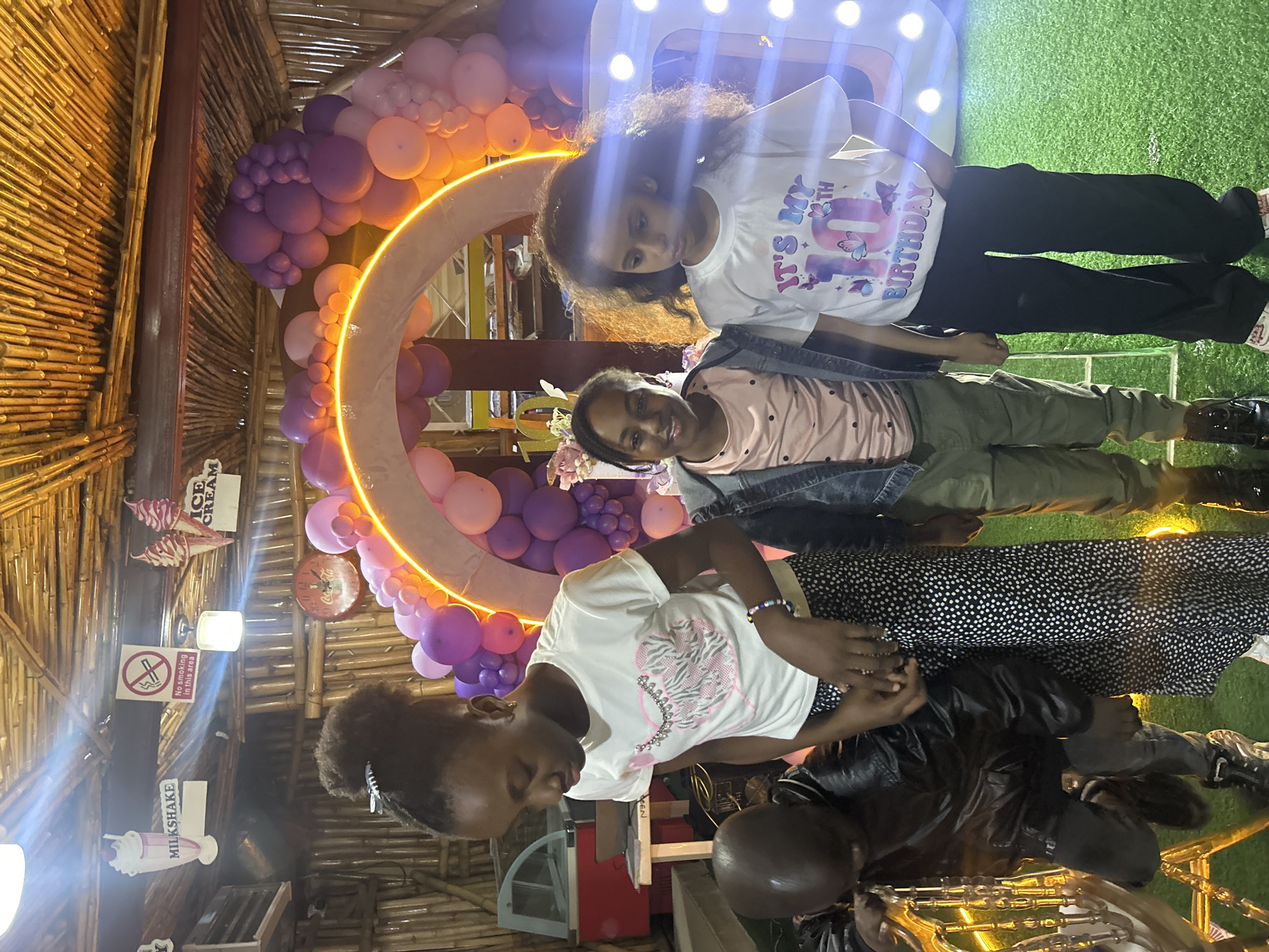 The image depicts an indoor celebration, possibly a birthday party, set in a location in Bar Jirgi Summa, Nigeria. The scene is decorated with an abundance of purple and pink balloons, forming an archway with a glowing orange outline. A large, illuminated number "10" is visible on the right side of the frame.
In the foreground, four children are present. A young boy, wearing a black leather jacket and dark pants, is leaning forward. To his right, a young girl with her hair in a bun is wearing a white t-shirt with a pink and silver design and black and white polka dot pants. She has a bracelet on her wrist. Next to her stands another girl in a denim jacket over a pink polka dot t-shirt, wearing olive green pants. On the far right, a girl with curly dark hair is wearing a white t-shirt with the text "IT'S MY 10TH BIRTHDAY" and butterflies, paired with black pants.
The environment features a thatched bamboo ceiling and bamboo walls. Signage indicating "MILKSHAKE" and "ICE CREAM" is visible on the ceiling, along with a "No smoking in this area" sign. A refrigerated display case, possibly for ice cream, is seen in the background. The floor is covered with artificial green grass. The lighting suggests an evening or indoor setting, with a combination of ambient and accent lighting.