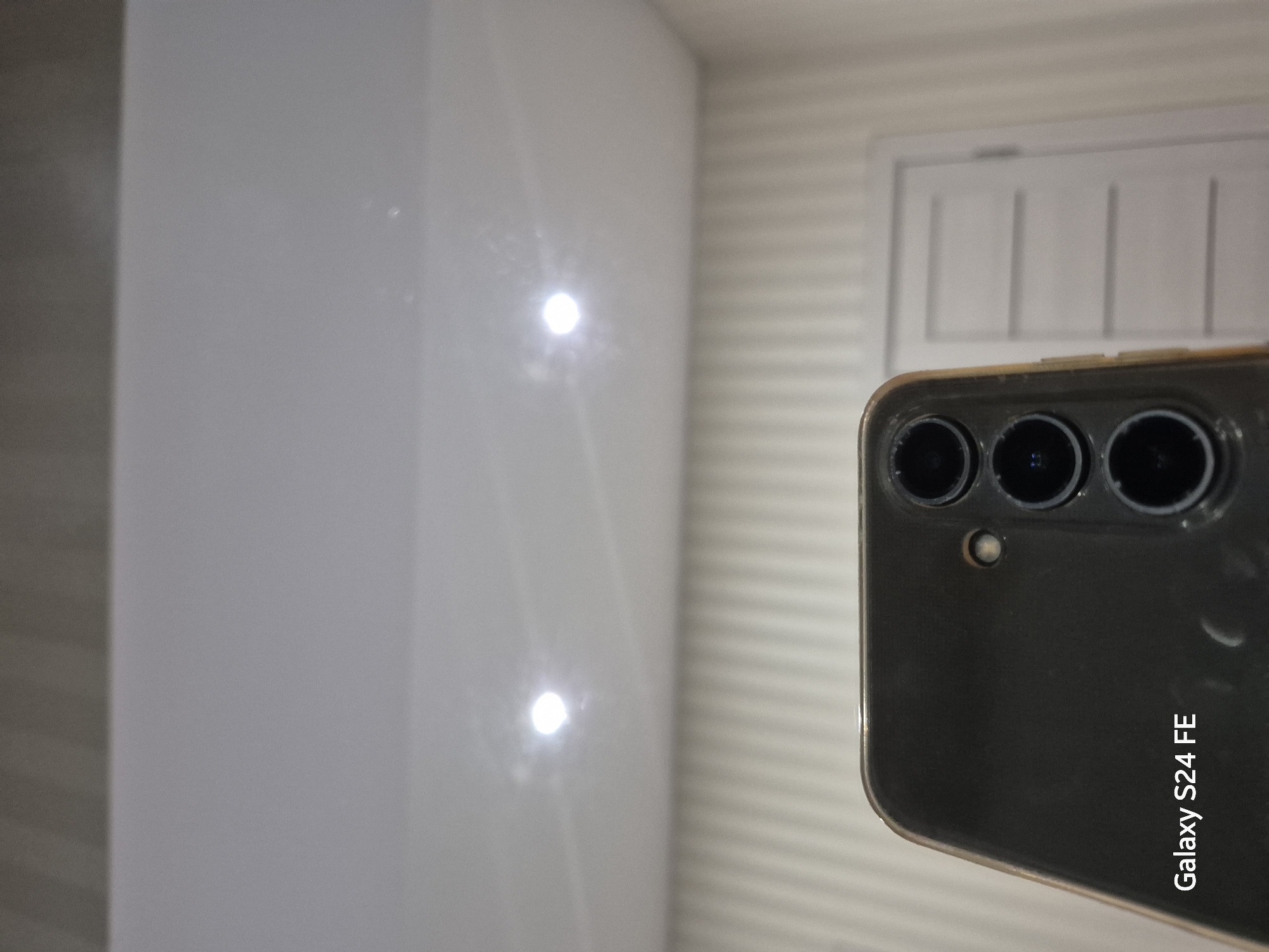 The image shows the rear camera array and the bottom left corner of a dark-colored smartphone, identified by the watermark "Galaxy S24 FE". The phone is positioned in the foreground, angled slightly upwards. The background consists of a white, vertically ribbed wall and a white door with horizontal paneling. Above the wall, a white ceiling with two circular light fixtures is visible. The lighting in the image suggests an indoor setting. There are no people or specific landmarks of Chișinău, Moldova visible.