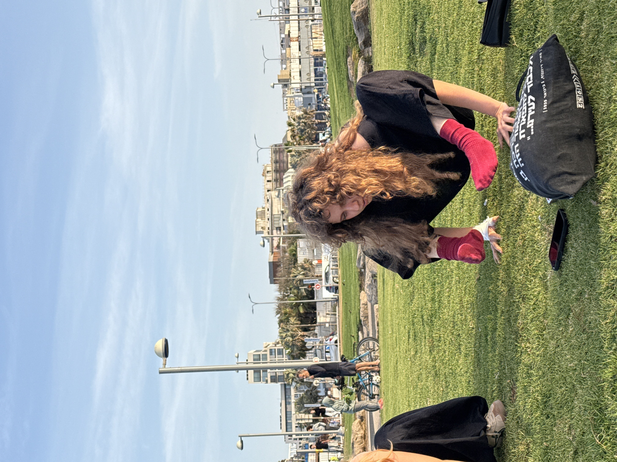 A person with curly hair, wearing a black garment and red socks, is seen bending over in a park, with their face obscured by their hair. A black bag with white Hebrew writing is near the person. Additional people are visible in the background, along with buildings, palm trees, and street lights along the waterfront. The location is likely a park along the coastline of Tel Aviv, Israel.
