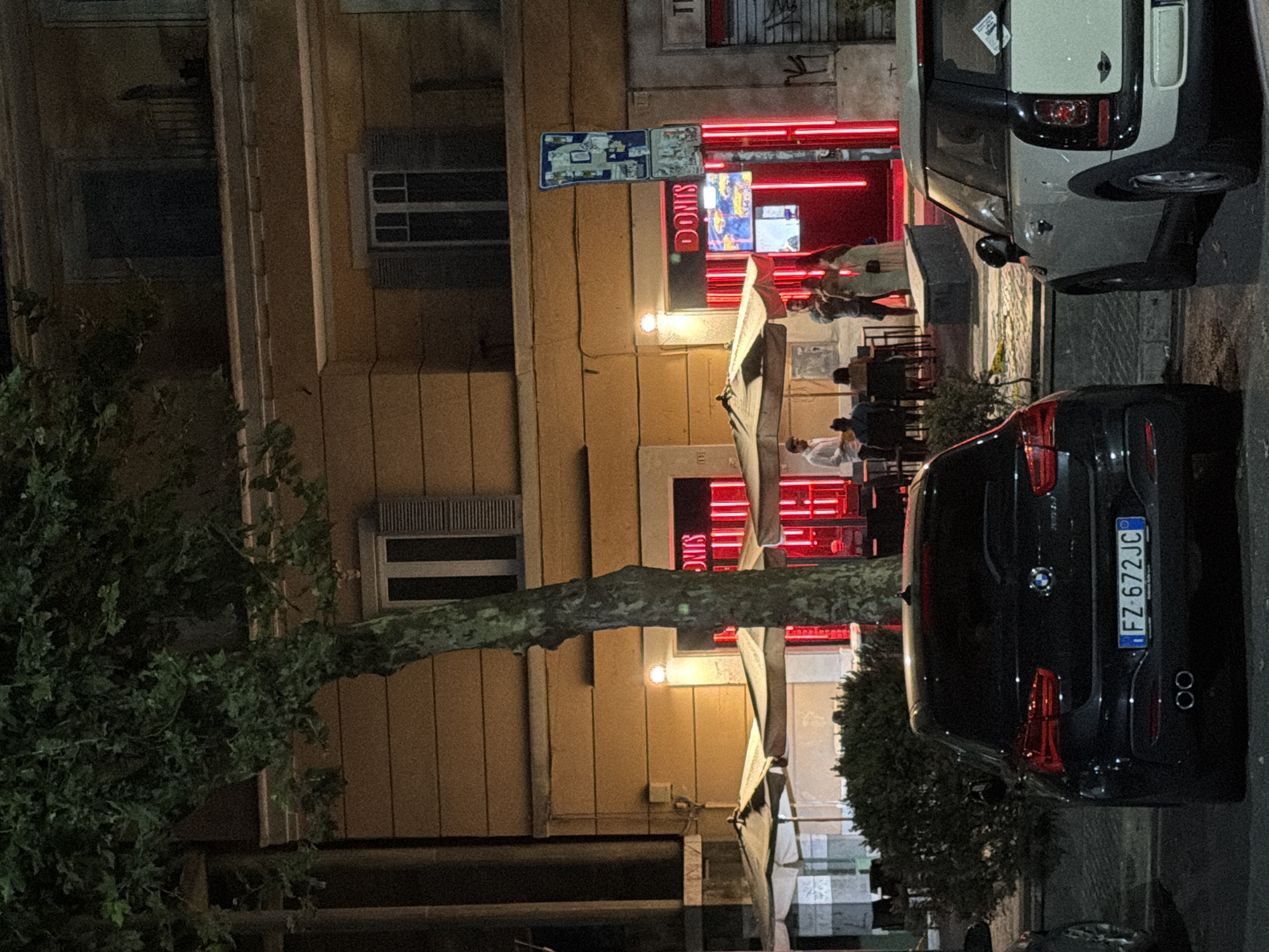 The image shows a street scene at night in Rome, Italy, featuring a storefront with neon lights, outdoor seating, and parked cars. The storefront has "DONIS" written in neon letters, and the interior is illuminated with red lights. Several people are visible outside the establishment, some seated at tables and others standing. Two cars are parked in the foreground: a black BMW in the left foreground and a white Mini Cooper in the right foreground. A tree with dense foliage stands to the left of the storefront, partially obscuring the building. The building itself appears to be a multi-story structure with windows. The street appears to be paved, and there are streetlights or building lights illuminating the scene. A blue traffic sign is mounted on a pole near the storefront.