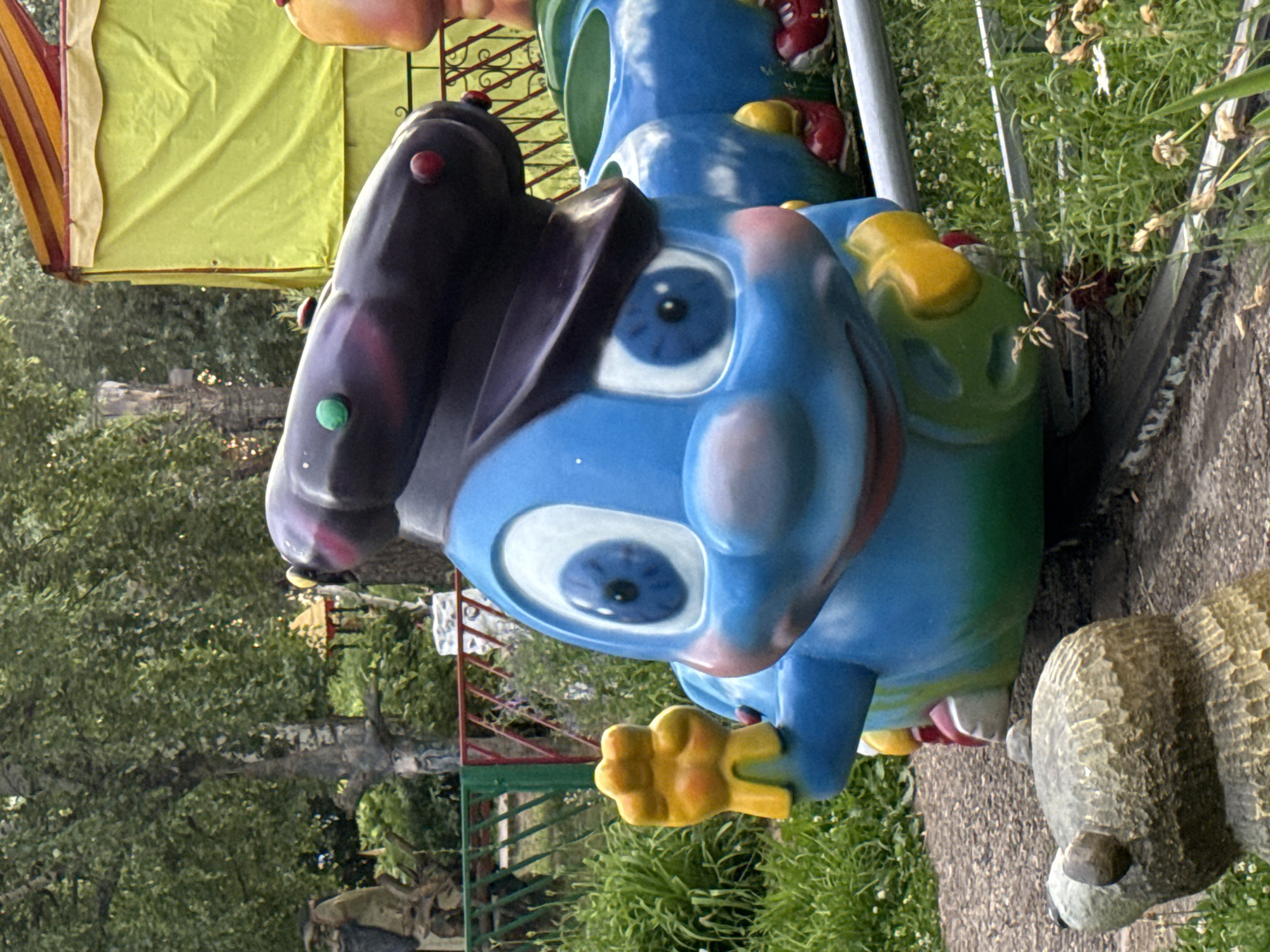 The media file shows a close-up of a brightly colored children's amusement park ride, oriented vertically. The ride is designed to look like a blue cartoon character with large, wide eyes, a purple hat, and yellow hand-like protrusions. The character's face is the primary subject, with its blue and white eyes taking up a significant portion of the frame. Red and green circular embellishments are visible on the character's head and body.
In the background, out of focus, are hints of the amusement park setting. A yellow and red striped awning or tent is visible on the left. Green foliage, including trees and grass, suggests an outdoor location. A metal fence with red and green accents can also be seen. To the right of the blue character, a textured, stone-like object, possibly another part of the ride or a decorative element, is visible. The lighting suggests it is daytime.
The overall scene evokes a playful and cheerful atmosphere associated with an amusement park. There are no people visible in the frame. No text is discernible. The location is Chaikovsky, Russia.