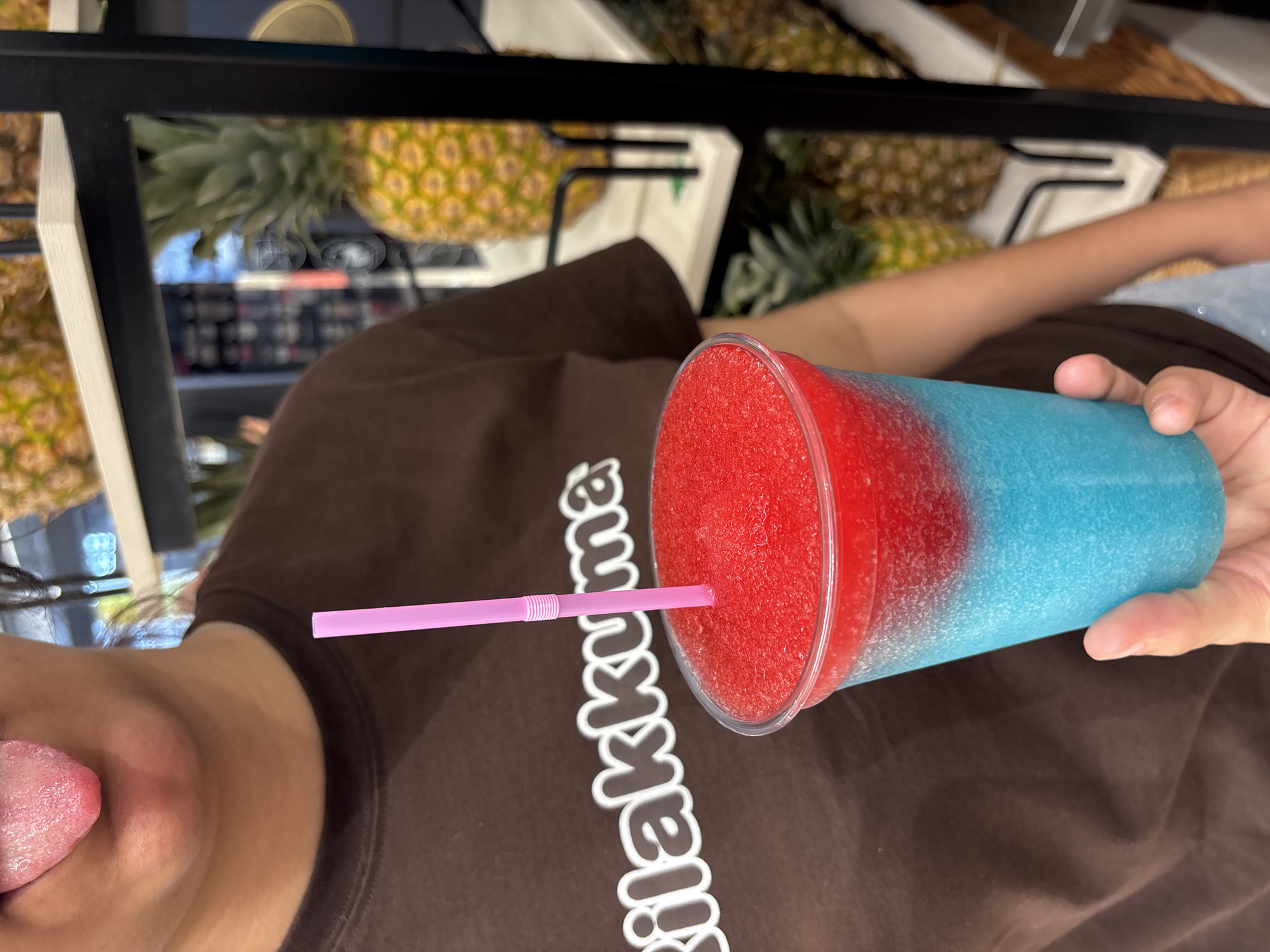 In an indoor setting, potentially a shop or cafe in Urbanización Villa Martín, Spain, a person is holding a clear plastic cup filled with a layered frozen beverage. The drink is divided into two distinct colors: a bright red on top and a vibrant blue on the bottom. A pink straw is inserted into the red layer. The person's torso is visible, wearing a brown t-shirt with white lettering that appears to read "kilakkuma". A portion of the person's face is visible at the top of the frame, with their tongue sticking out. In the background, shelves display several pineapples. The lighting appears to be bright and natural or well-lit artificial.