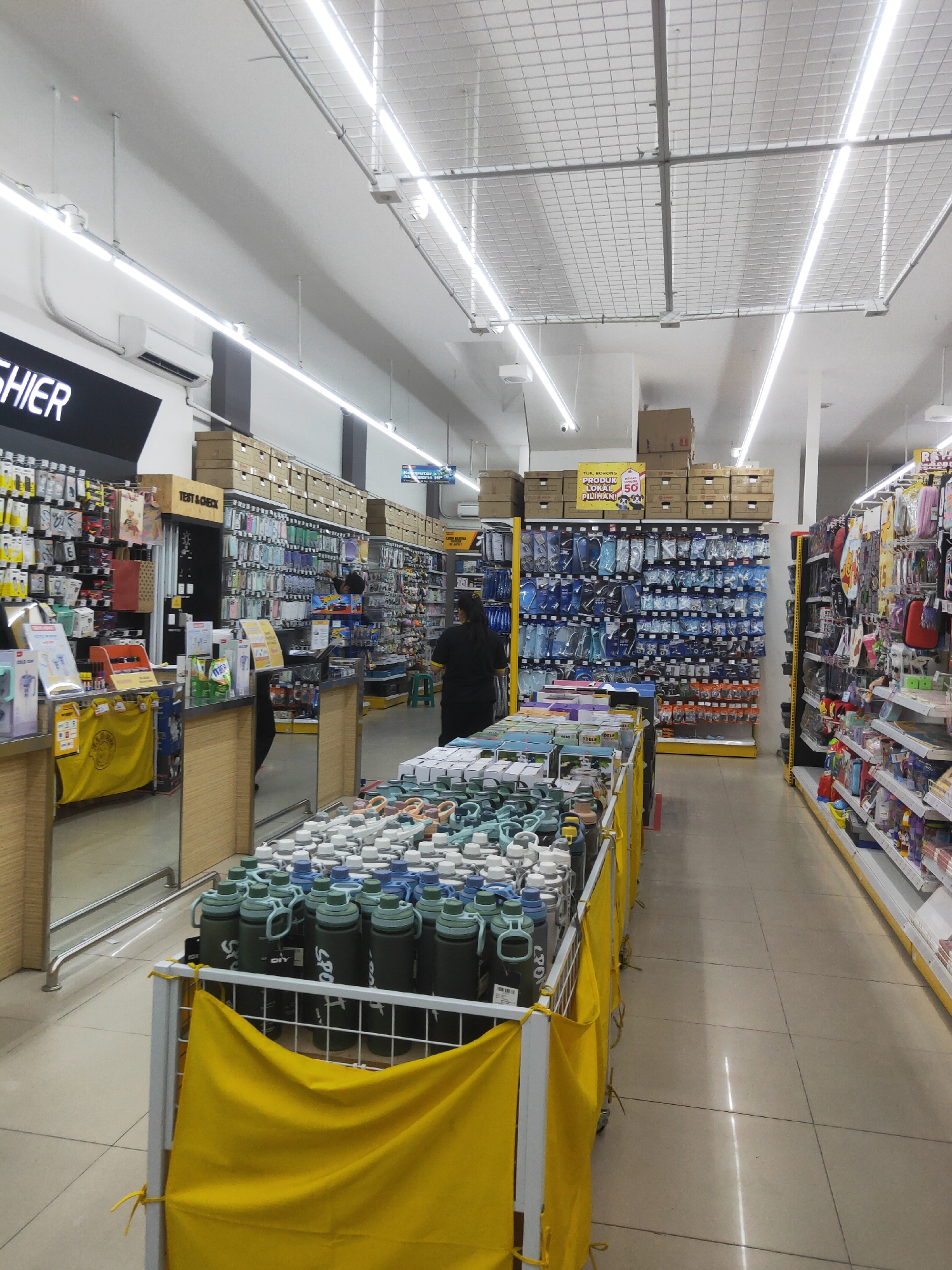 The image captures an aisle within a brightly lit retail store, likely in Sintang, Indonesia, given the context. The aisle is lined with shelves displaying a variety of merchandise. On the left, a counter area is visible with a yellow fabric covering some products. Behind the counter, shelves are stocked with small packaged items, many appearing to be electronic accessories. Further down the aisle, more shelves are filled with a colorful assortment of items, including what appear to be phone cases and other accessories. In the center of the image, a person in dark clothing is standing near a display of various types of water bottles, some of which are prominently featured in the foreground. A sign with Indonesian text is visible on a shelf above the water bottles, advertising a sale. On the right side of the aisle, more shelves are packed with merchandise, and another person in a dark uniform can be seen walking away from the camera. The flooring is tiled in a light color. The store features overhead lighting and a grid-like ceiling structure.