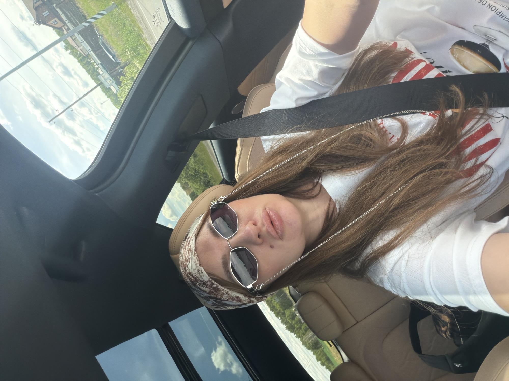 A woman with long brown hair is sitting in the passenger seat of a car, looking at the camera. She is wearing a white t-shirt, sunglasses, and a patterned headscarf. The car has a sunroof open, revealing a cloudy sky and a suburban landscape with houses, trees, and power lines. The interior of the car features tan leather seats and a black seatbelt. A black bag with a leopard-print lining is visible on the seat next to her. The scene appears to be taking place in Malinovka, Russia, given the context.