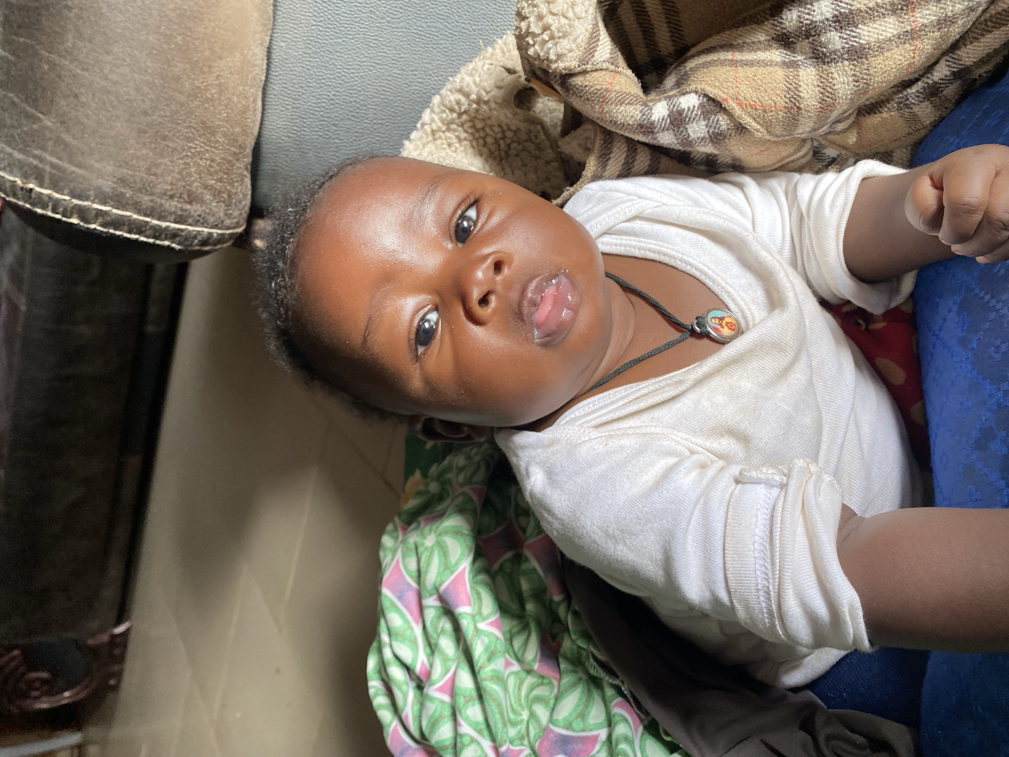 An adorable infant with dark skin and dark, curly hair is prominently featured, lying down and looking directly into the camera. The baby wears a simple white long-sleeved onesie or shirt and a black cord necklace with a small, round pendant depicting two figures, possibly religious icons. A small amount of white substance, likely milk or drool, is visible on the baby's lower lip and chin, adding to its innocent appearance. The baby's eyes are wide and bright, conveying a calm and curious expression.
The baby is surrounded by a mix of textures and fabrics. To the left, a section of a dark brown or greyish-brown upholstered surface, possibly a sofa arm or cushion, is visible. Above and to the right, a cozy beige and brown plaid blanket with a visible fluffy, sherpa-like lining provides warmth. Below the baby, a vibrant green and pink patterned fabric, possibly another blanket or sheet, is partially visible. A patch of blue fabric, likely clothing, is also seen on the far right. The scene suggests an indoor setting, perhaps a comfortable home environment in Vom, Nigeria. There is no visible text in the image.