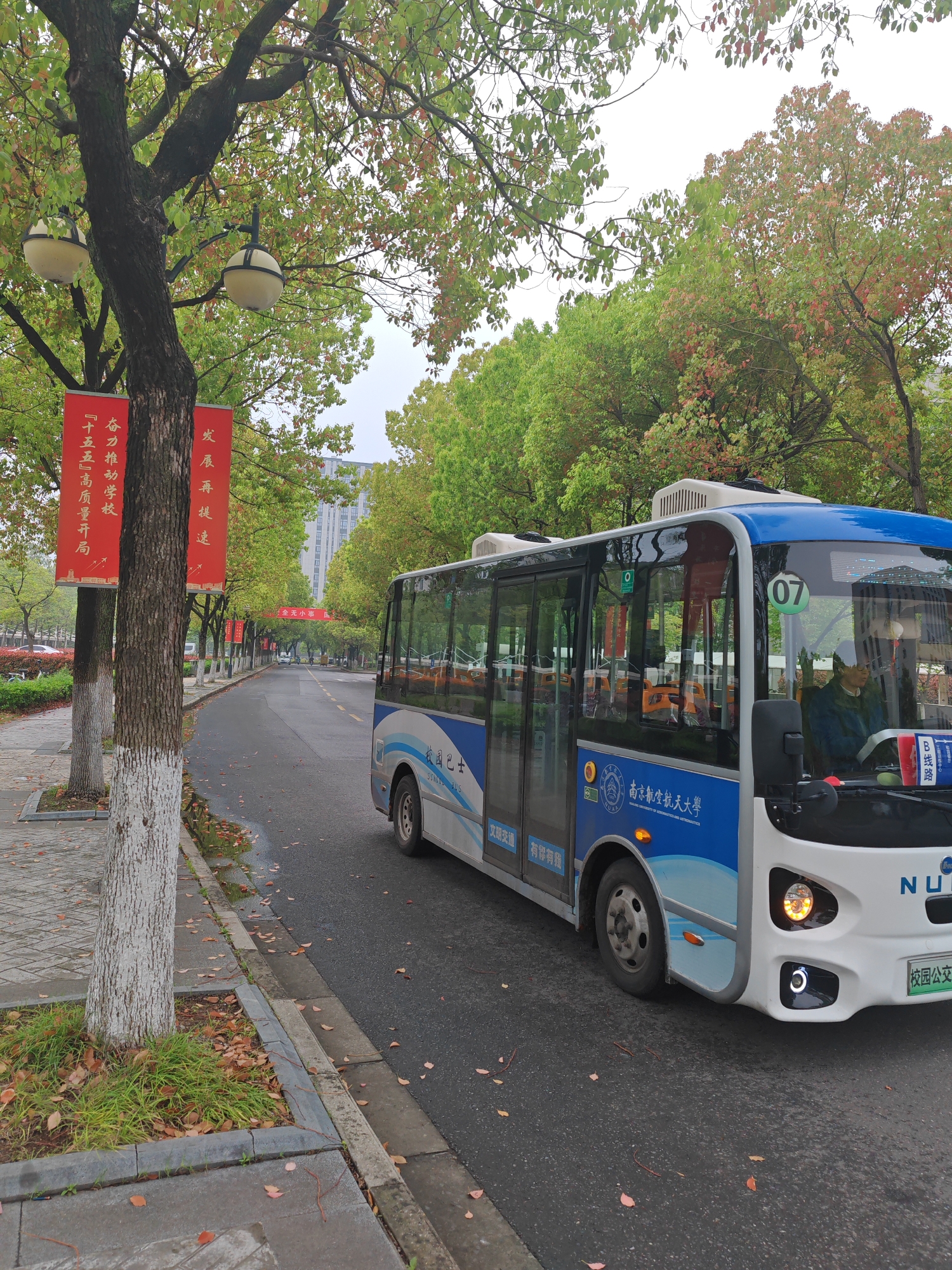 The image shows a white and blue "NUA" branded bus, number 07, traveling on a street in what appears to be the Jiangning District of Nanjing, China. The bus features the Chinese characters for "校园巴士" (School Bus) and branding related to the Nanjing University of Aeronautics and Astronautics. A driver is visible inside the bus. The street is flanked by trees, some with red banners bearing Chinese characters. The weather appears overcast, and there are leaves scattered on the pavement. A lamppost is visible on the left side of the image.