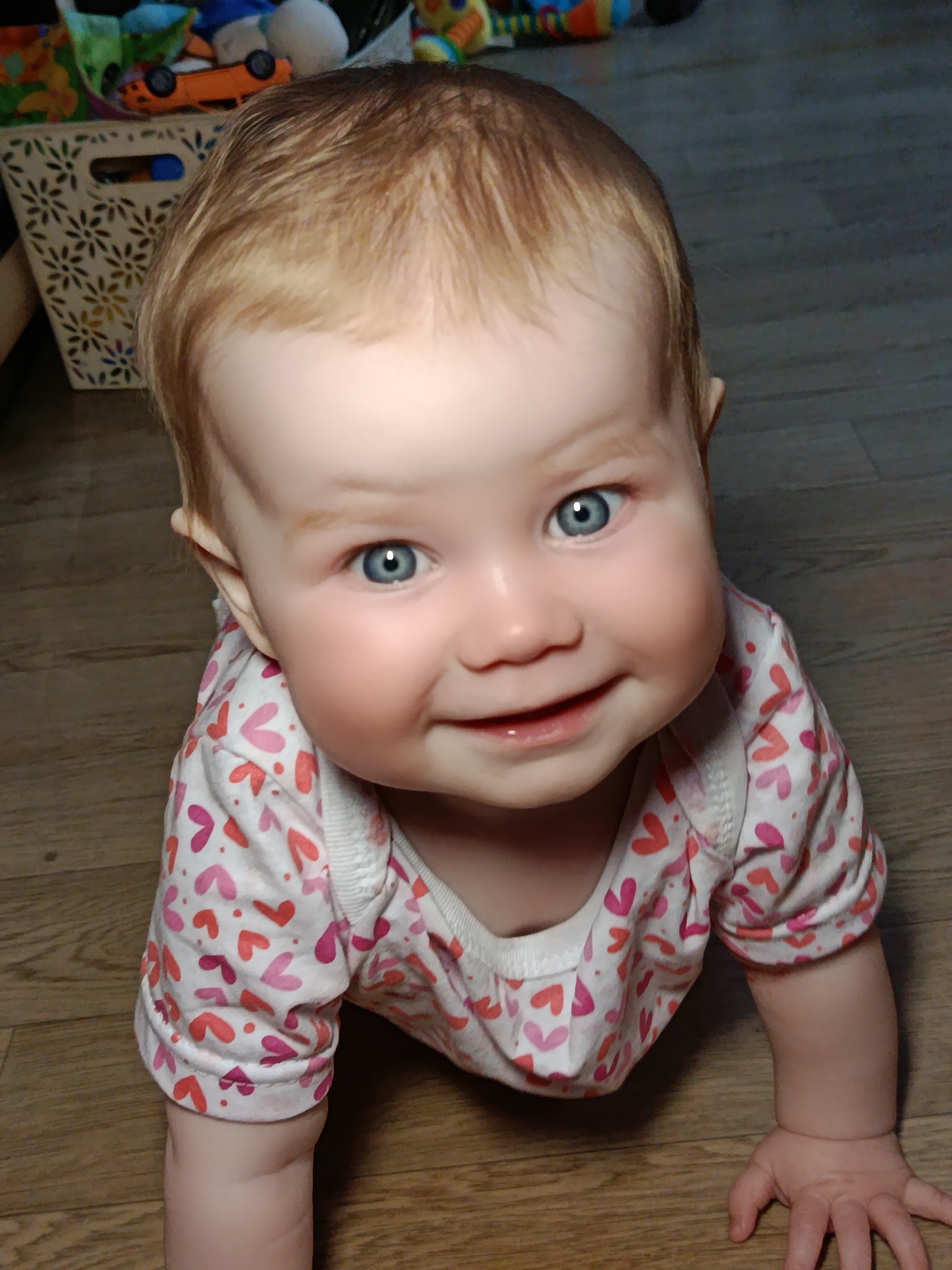 A close-up, high-angle portrait features a smiling infant with fair skin, light reddish-blonde hair, and bright blue eyes, positioned on a light brown wooden floor. The baby looks directly at the camera with a broad, joyful smile, revealing some lower gums or emerging teeth. Chubby cheeks and wide, engaged eyes convey happiness and curiosity. The infant is wearing a light-colored, long-sleeved shirt patterned with numerous red and pink hearts, and appears to be in a crawling stance, with hands on the floor.
In the blurred background to the upper left, a light-colored basket with a decorative, cut-out floral or starburst pattern is visible. Inside or near the basket, various colorful toys are indistinctly seen, including a bright orange toy car and other soft, fabric-like items. This suggests an indoor home environment, likely a play area. The lighting is soft and even, indicative of an indoor daytime setting. No visible text is present in the image. The scene is set in Фоки, Russia.