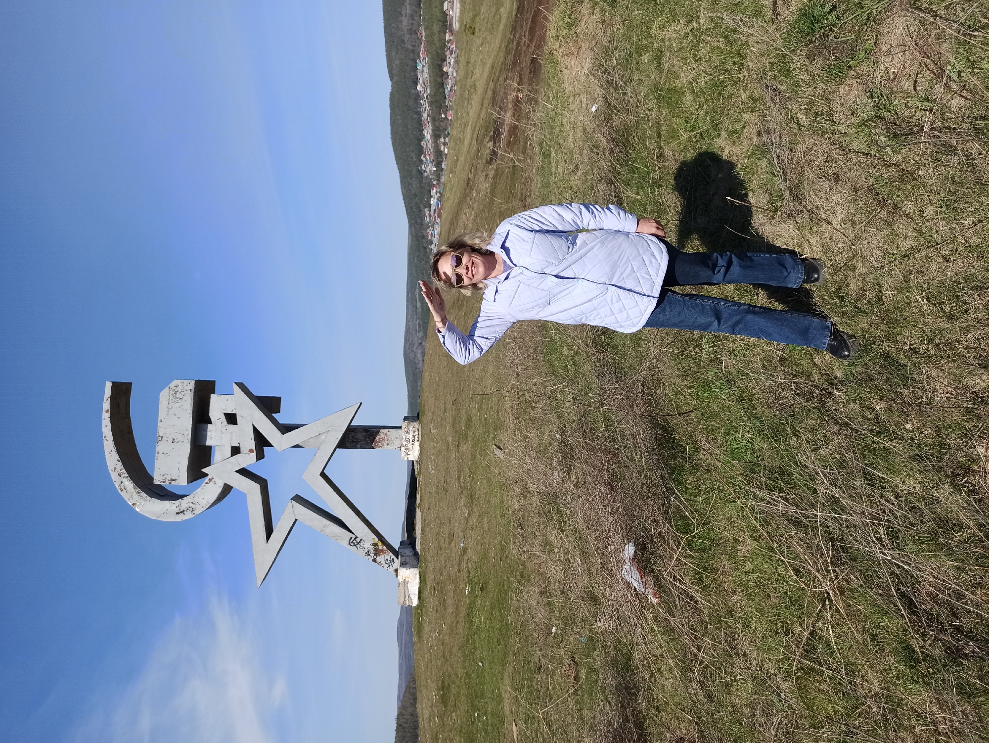 The image shows a landscape with a woman posing next to a large monument in Сатка, Russia. The monument consists of a Cyrillic letter "U" and a five-pointed star, painted in a metallic gray color. The woman is wearing sunglasses, a light blue jacket, and jeans. She is raising her hand in a waving gesture. The background includes a blue sky and a hillside with a town visible in the distance. The ground is covered in grass and dry vegetation. The image appears to be taken on a sunny day.