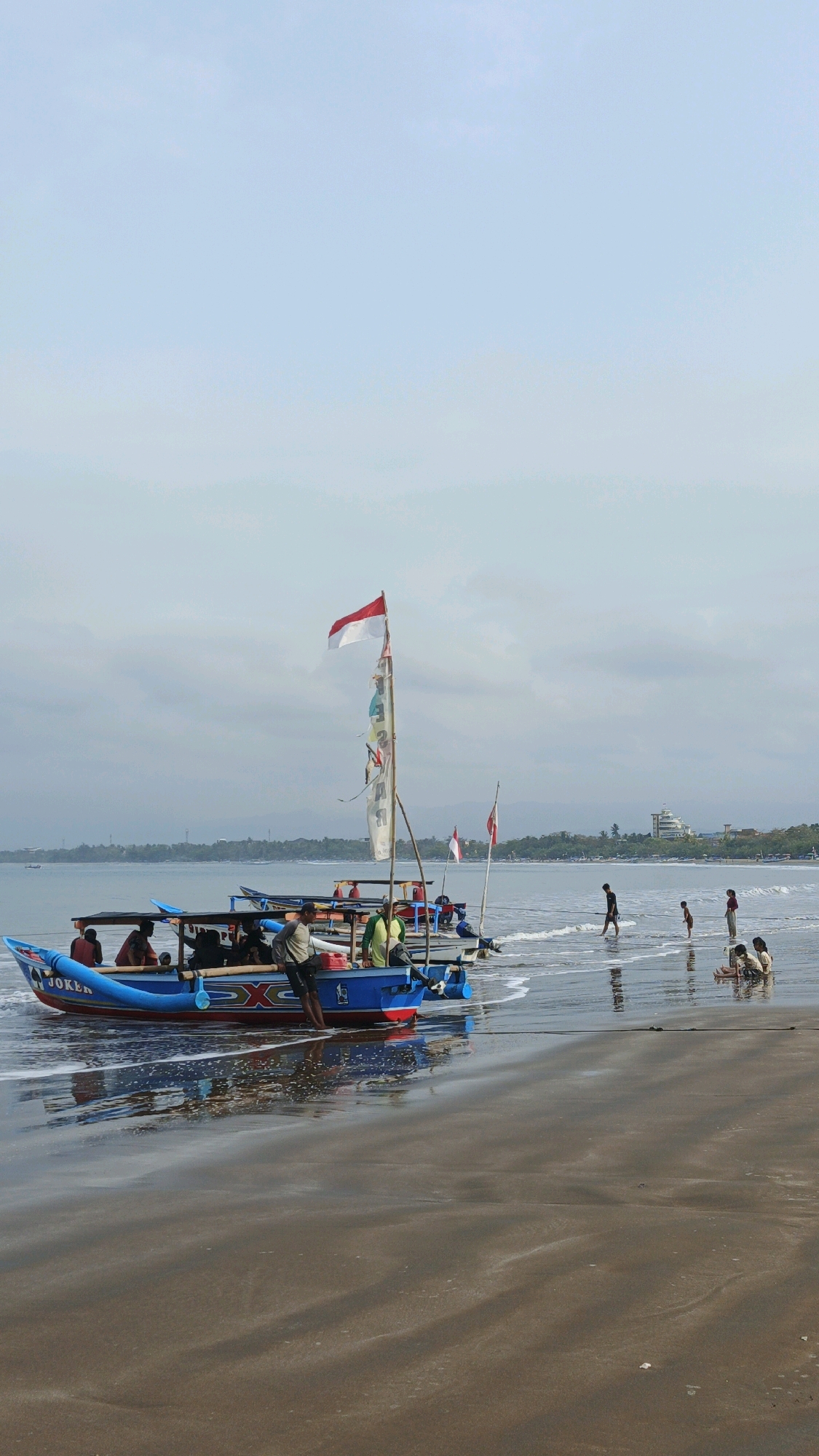 The image captures a serene scene at the beach in Desa Pangandaran, Indonesia. In the foreground, a brown sandy beach meets the gentle waves of the ocean. The water is shallow and reflects the overcast sky.
Dominating the left side of the image is a vibrant blue and red fishing boat named "JOKER". Several people are seated and standing within the boat, some appearing to be passengers and others crew. A prominent red and white flag, likely the Indonesian flag, flies from a mast on the boat, alongside other smaller pennants.
To the right of the boat, in the shallow water, a group of children are playing. They are spread out, some standing and walking, while others are seated or kneeling by the water's edge.
In the background, the ocean stretches towards a distant shoreline lined with trees and buildings. A large, modern-looking building with a light-colored facade is visible further down the coast. The sky is a pale, hazy blue with scattered clouds, suggesting a soft, diffused light. The overall atmosphere is peaceful and tranquil, depicting a typical day at the seaside in this Indonesian village.