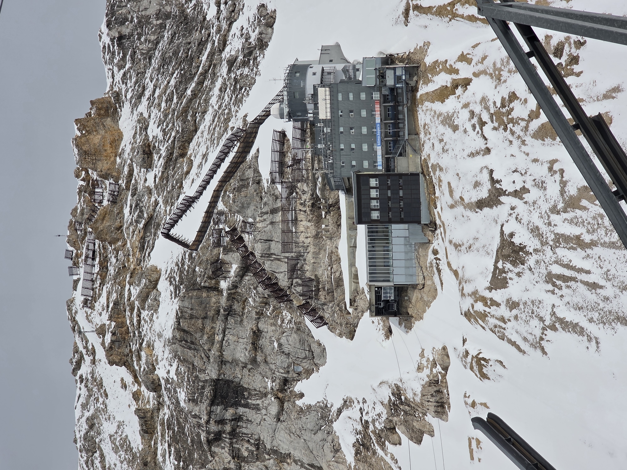 The image presents a detailed view of a multi-level building complex integrated into the snowy, rocky slopes of a high-altitude mountain. The location is the Zugspitze summit area, Bavaria, Germany. The main structure comprises several interconnected sections: a dark grey, multi-story building featuring numerous windows and a visible banner displaying "klima.bayern"; an adjacent modern section with extensive glass paneling, likely a cable car station; and a cylindrical structure topped with a white dome, indicative of an observatory or meteorological station. Ascending the steep mountain face above and to the right of the buildings are multiple segments of zig-zagging dark wooden or metal fence-like barriers, which appear to function as avalanche protection or marked pathways. The terrain consists of exposed grey rock interspersed with significant patches of white snow, suggesting an alpine environment. The sky is uniformly overcast and grey. In the lower foreground, partial views of dark metal structural elements are visible, possibly belonging to a viewing platform or cable car infrastructure. No individuals are discernible in the scene.