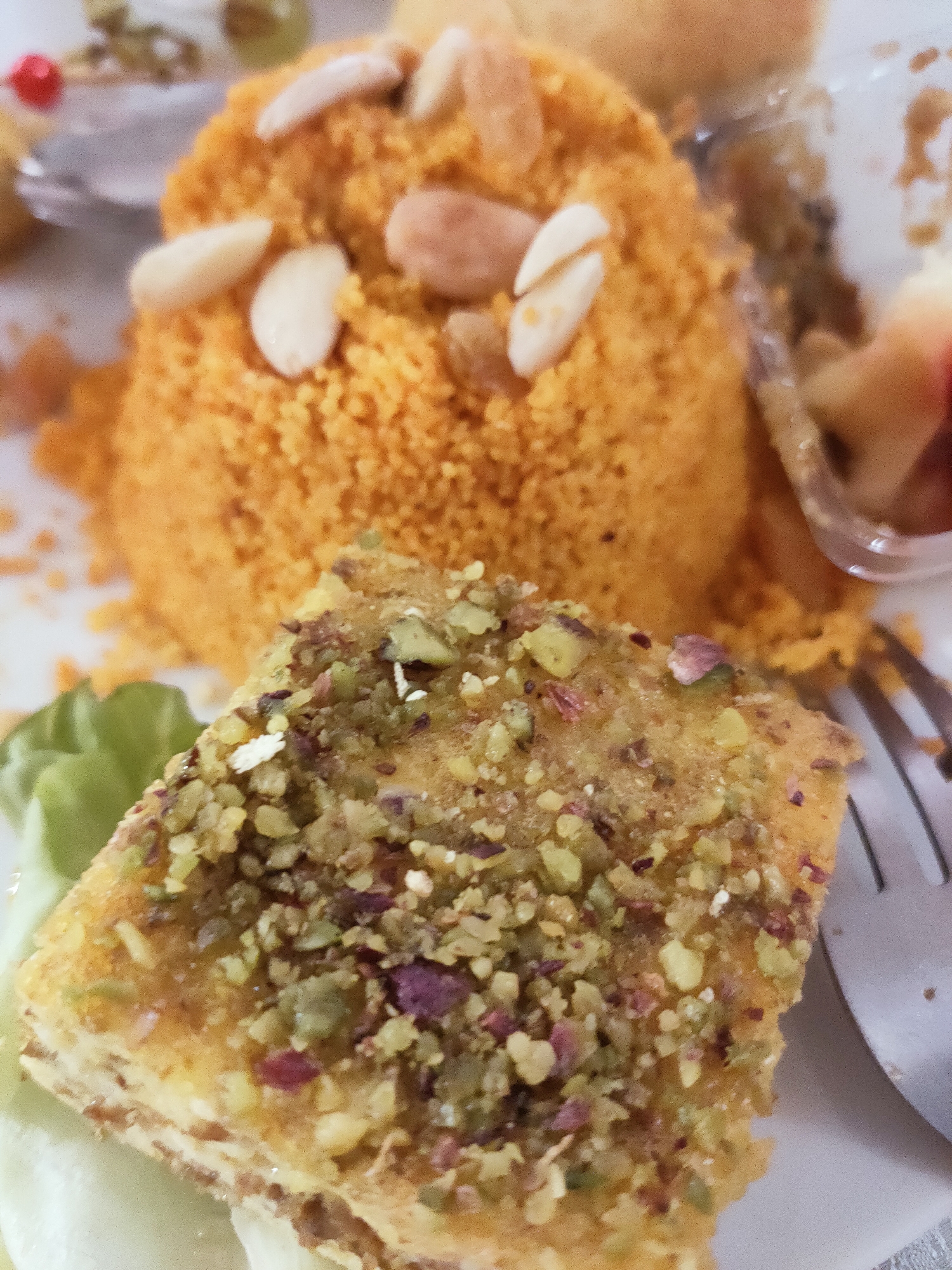 This is a close-up, eye-level shot of a dessert platter served on a white plate. The dish features two main components: a mound of orange, crumbly dessert topped with slivered almonds, and a rectangular piece of pastry covered in chopped pistachios and other crushed nuts. The pastry appears layered, with visible strata. A small sprig of green lettuce is positioned to the left of the pastry. In the background, out of focus, are glimpses of other food items and what appears to be a clear plastic container with fruit. A silver fork is partially visible on the right side of the frame, resting on the plate. The lighting is bright, highlighting the textures and colors of the food. The image is a still life focused entirely on the culinary presentation.