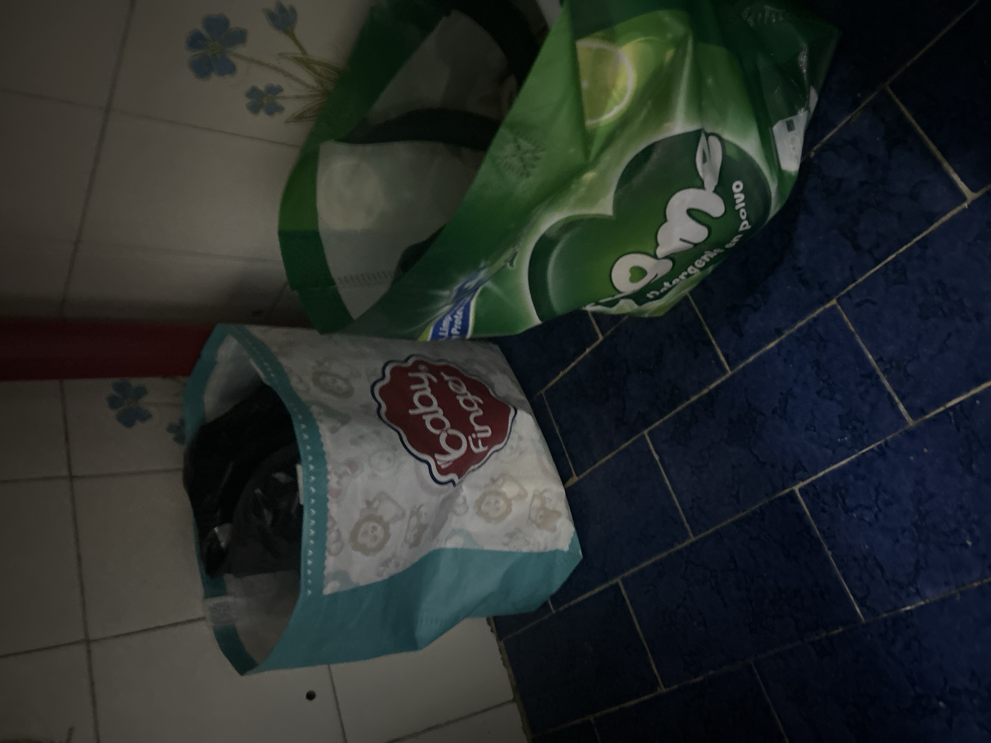 The image shows a corner in a room, likely a laundry area. There are two open bags on the floor. One bag is green and white, labeled with the brand "OM" detergent. The other bag is light blue and white, with a logo that says "Baby Finger". The floor is tiled with blue square tiles on one side and white square tiles on the other. The location is unspecified, and there is no information about people, actions, or interactions. The image is a still photograph of a domestic interior setting.