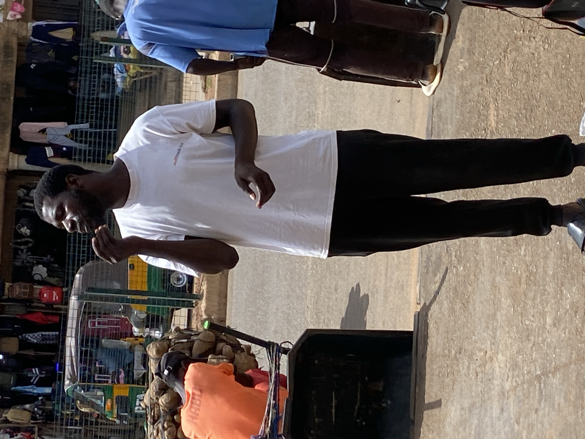 This image depicts an outdoor street or market scene in Bukuru, Nigeria, captured during daylight with clear, sunny weather as indicated by strong shadows. The ground is a light-colored, possibly paved surface.
In the prominent foreground, a man with dark skin, a beard, and short dark hair stands, facing towards the left side of the frame. He is dressed in a plain white short-sleeved t-shirt, which has a small, indistinct logo or text on its left chest area, and black trousers. His hands are clasped together at about chest height.
Behind this man, slightly to the right, another individual is partially visible, wearing a light blue short-sleeved top and dark lower garments. In the background, on the left, are market stalls featuring various clothes, such as shirts and trousers, displayed on metal racks. Further back, a green and yellow structure or vehicle can be seen.
In the lower right portion of the foreground, another person is seen bent over a dark-colored cart or container. This individual is wearing an orange hoodie with the word "HABU" prominently displayed in white capital letters. The cart holds several bundled items, possibly produce like coconuts, suggesting a vendor's setup. Strings or ropes are also visible hanging from the side of the cart.
The overall scene conveys a lively, informal commercial environment typical of a street market, with people engaged in daily work or moving through the area.