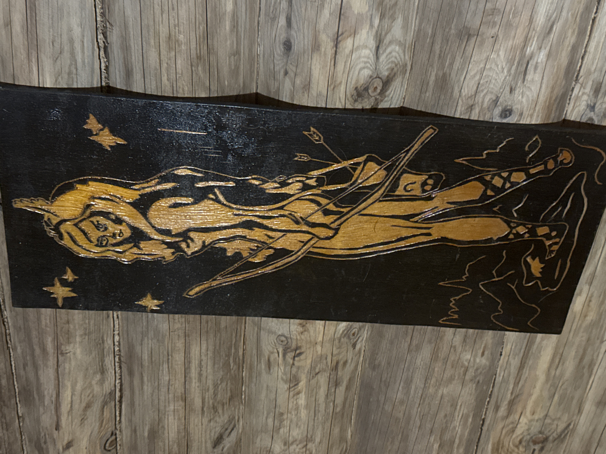 A rectangular, black wood carving of a figure is propped against a wooden wall. The carved image depicts a figure with long hair, holding a bow and arrow. Other elements in the carving include a bird, stars, and other unidentifiable forms. The location is unknown and not within the city of Маза, Russia.