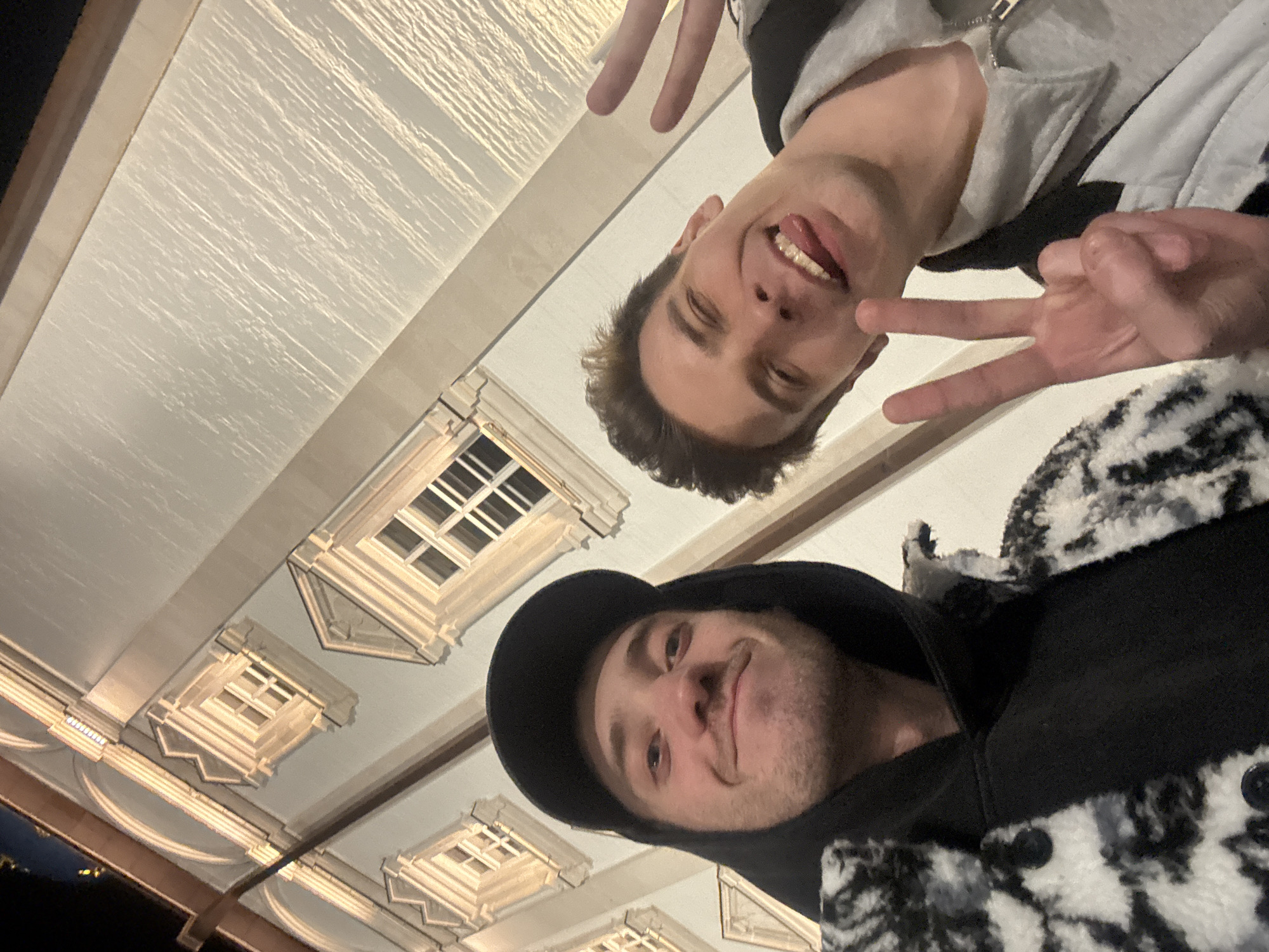 Two men are posing for a selfie at night, likely in Tver, Russia. The background reveals architectural details typical of the city's historical buildings. The men are positioned in the foreground with the architecture behind them. One man is wearing a baseball cap and a black and white jacket. The other man is wearing a gray hoodie. They are both making peace signs with their hands.