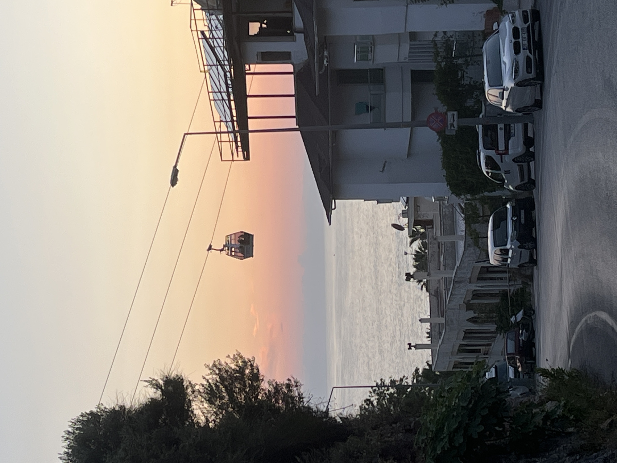 The image captures a cityscape in Alanya, Turkey, seen at an angle. A cable car is suspended mid-air, connected by wires to a metal structure on the left side of the frame. The background displays a body of water, likely the sea, with a soft, orange-toned sky above. Buildings are visible along the right side, including multi-story structures and residential houses. Several parked cars are aligned along the road. Vegetation, including trees and shrubs, fills the lower portion of the image. A street lamp is present near the cable car structure. The image composition suggests a viewpoint from a slightly elevated position.