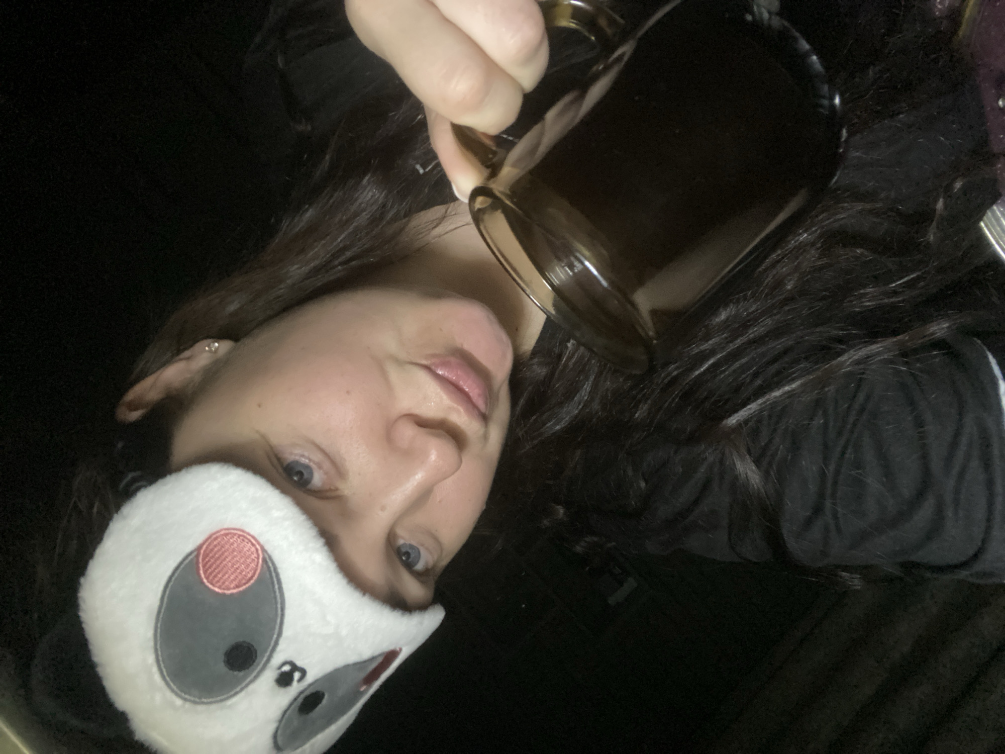 A woman wearing a sleep mask is holding a glass mug filled with a dark liquid, possibly coffee or tea. She is looking towards the camera. The background is dark. There is no information available about the location being Sochi, Russia.