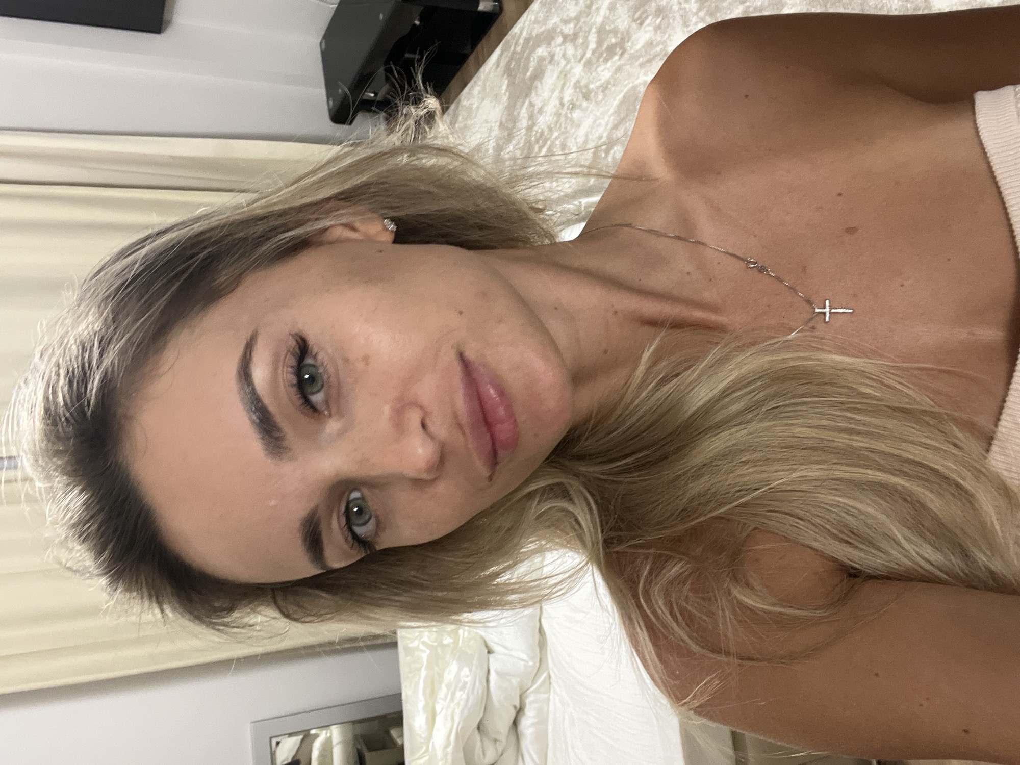 A close-up, waist-up, selfie-style photograph features a blonde woman with green eyes and fair skin. She is wearing a silver cross necklace. The background shows a bed with white linens and cream-colored curtains, suggesting an indoor domestic setting. The lighting is soft and natural. The image does not provide any specific details or context that would link it to the city of Sharm El Sheikh, Egypt.