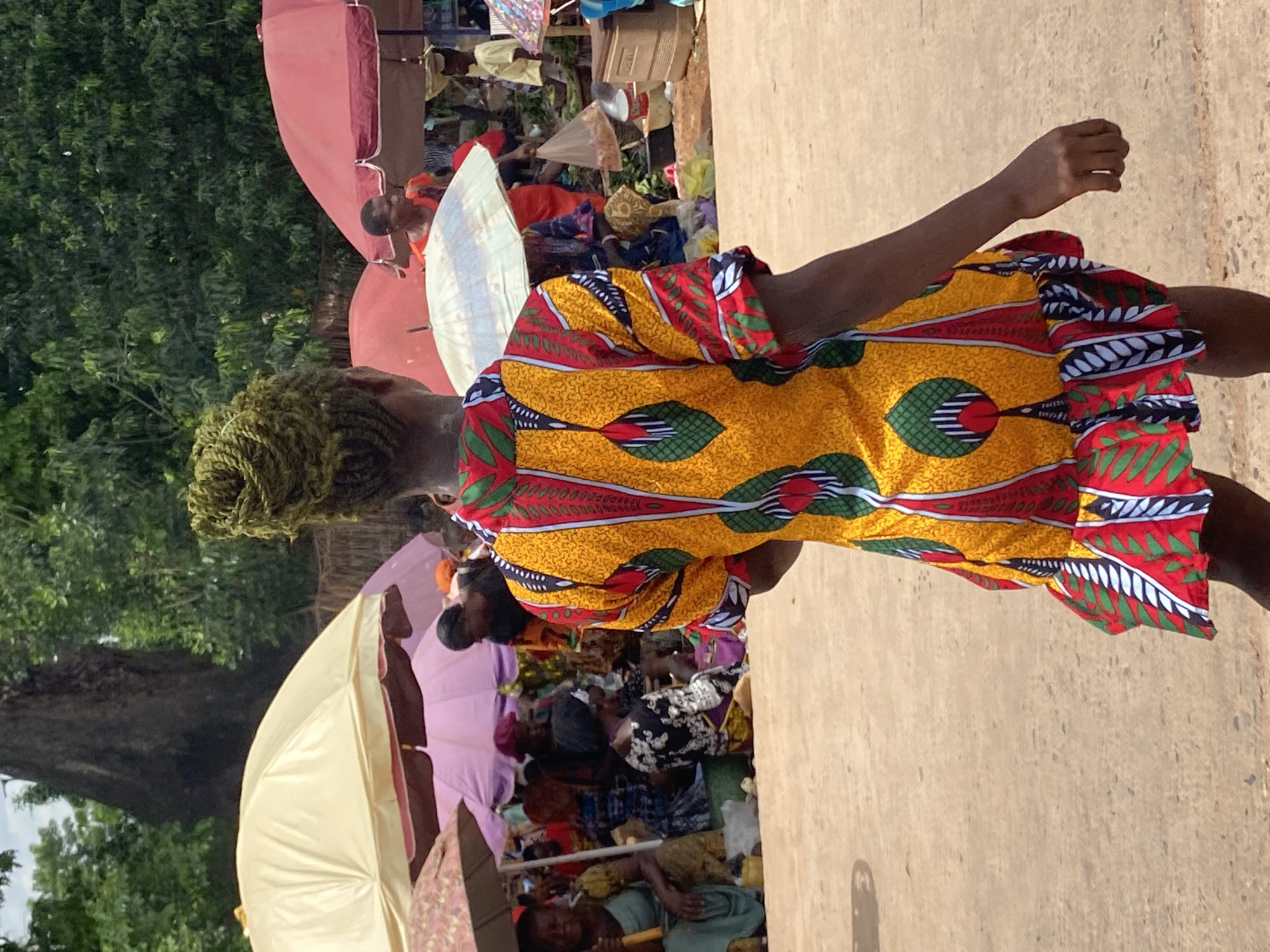 In Nguzu Edda, Nigeria, a market scene unfolds. A person with braided hair, dressed in a vibrant yellow and patterned garment with red and green accents, stands facing away from the viewer. They are positioned on a concrete or paved surface. Behind them is a bustling market, with vendors under colorful umbrellas, including pink and white ones. The market activity is partially obscured by foliage from nearby trees. Other individuals are present in the market, some sitting and appearing to be selling goods.