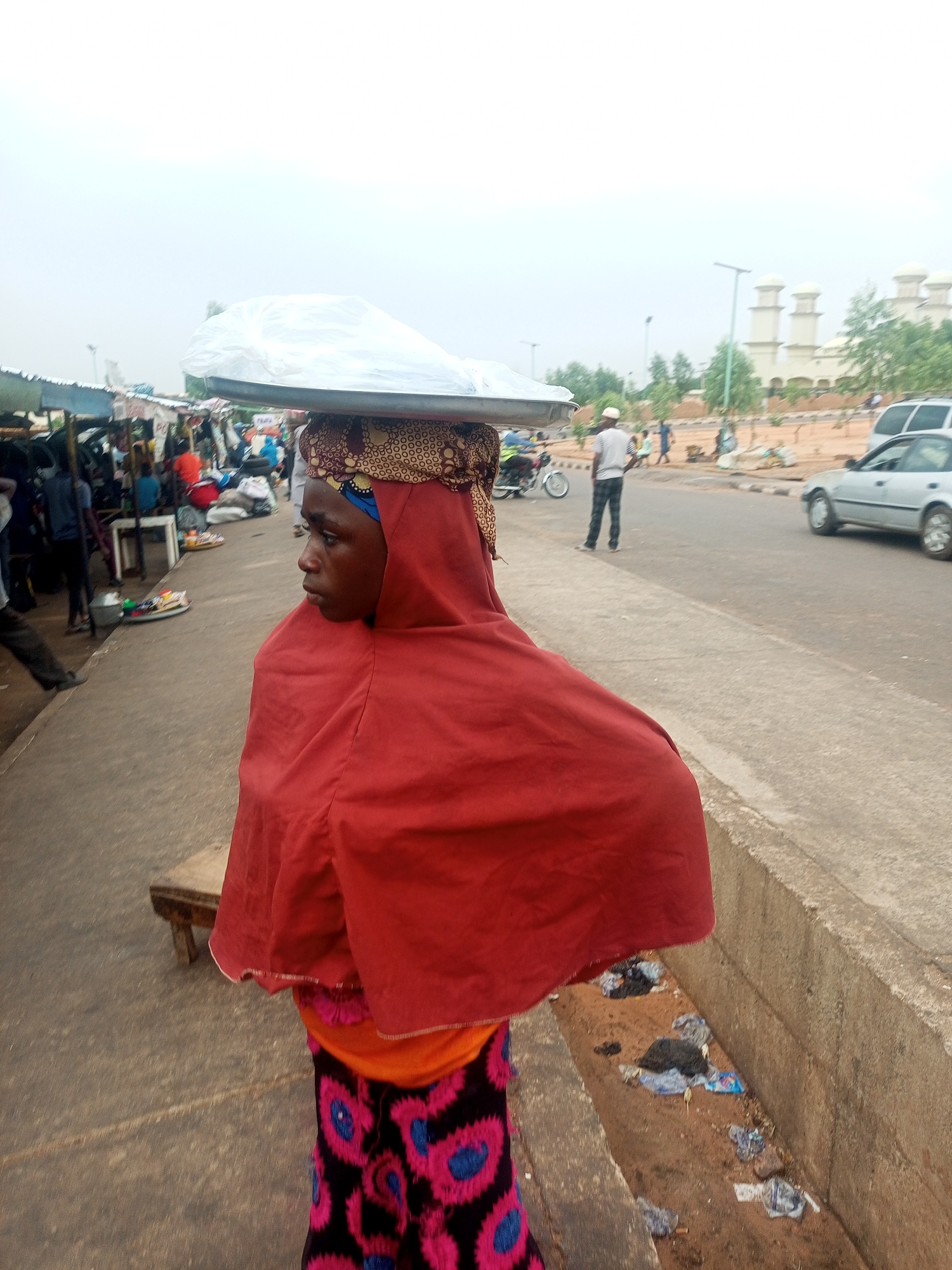 A young woman wearing a red hijab and a patterned skirt balances a silver tray, covered in a plastic bag, on her head, walking on a concrete sidewalk next to a street in Gombe, Nigeria. A narrow drainage ditch runs beside the sidewalk, littered with trash. A bustling market is visible on the left side of the image, filled with vendors and customers under makeshift stalls. Across the street, there's a vehicle, a person walking, a motorcycle, and a building with distinctive white domes.