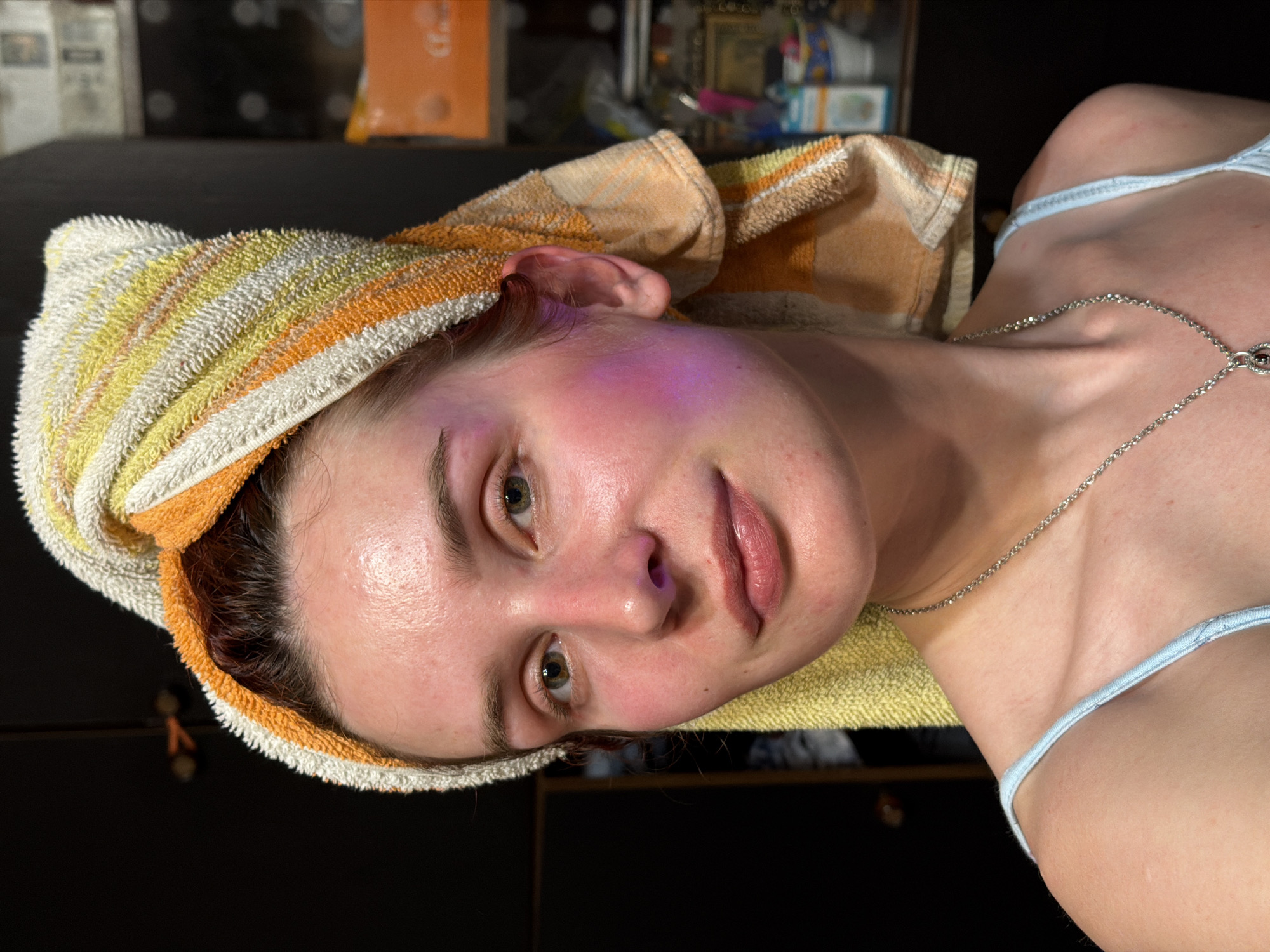 The image presents a close-up, rotated portrait of a young person, likely female, captured indoors in Заболотово, Russia. Their head is wrapped in a striped towel, predominantly white, yellow, and orange, suggesting recently washed hair or post-shower activity. The individual looks directly into the camera with a neutral expression.
A distinctive feature is a vibrant pinkish-purple glow visible on the left side of their face, encompassing the cheek and temple area, and subtly inside the left nostril. Their skin appears dewy or moist. They have green or hazel eyes and naturally colored, slightly parted lips. Around their neck, a delicate silver chain necklace is visible. The person is wearing a light blue strap from what appears to be a camisole or similar top. The background is dark, out of focus, and suggests an indoor setting with indistinct objects or shelving in the dimly lit environment. No visible text is present.