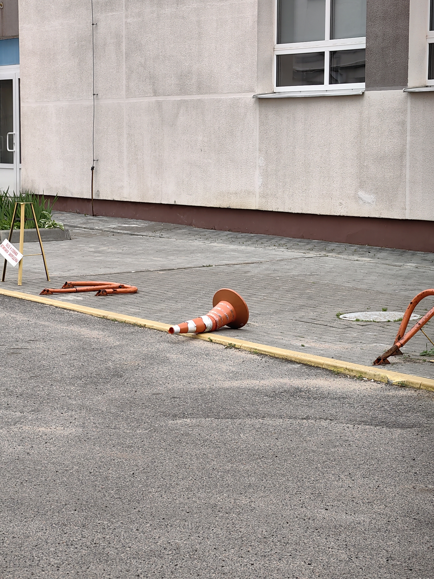 The image depicts an outdoor scene in a city, likely Minsk, Belarus, characterized by a light-colored building with windows and a textured facade. In the foreground, an asphalt surface meets a paved walkway bordered by a yellow curb. Several orange traffic cones are present. One traffic cone is lying on its side on the paved walkway, with its top pointing towards the right of the frame. Another orange traffic cone, lying on its side, is partially visible in the lower right corner of the image. Additionally, a collapsed orange barrier is lying on the paved walkway in the mid-ground. A small white sign with text, supported by a yellow A-frame stand, is positioned on the paved walkway to the left of the barrier. Green foliage is visible behind the sign. The overall scene suggests a temporary disruption or construction area.