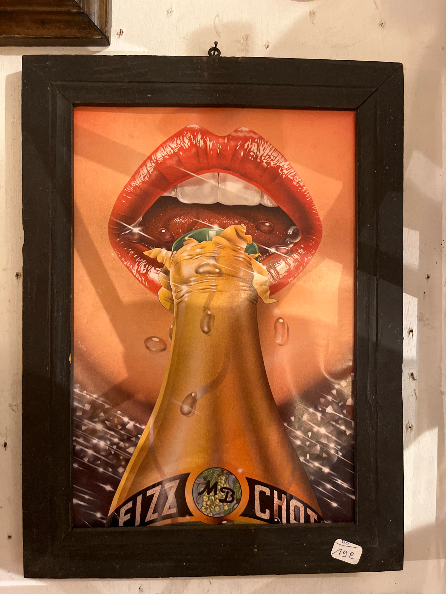 The image shows a framed advertisement for "FIZZ CHOT" featuring a close-up of red, glossy lips opening to receive the neck of a champagne bottle, with liquid appearing to pour out. The advertisement is hanging on a white, textured wall in what appears to be a shop or gallery in Yvoire, France, as indicated by a sticker with "Yvoire" and a price tag of "19€" on the bottom right of the frame. The advertisement has a vintage aesthetic, with sparkling effects and a warm, orange background.